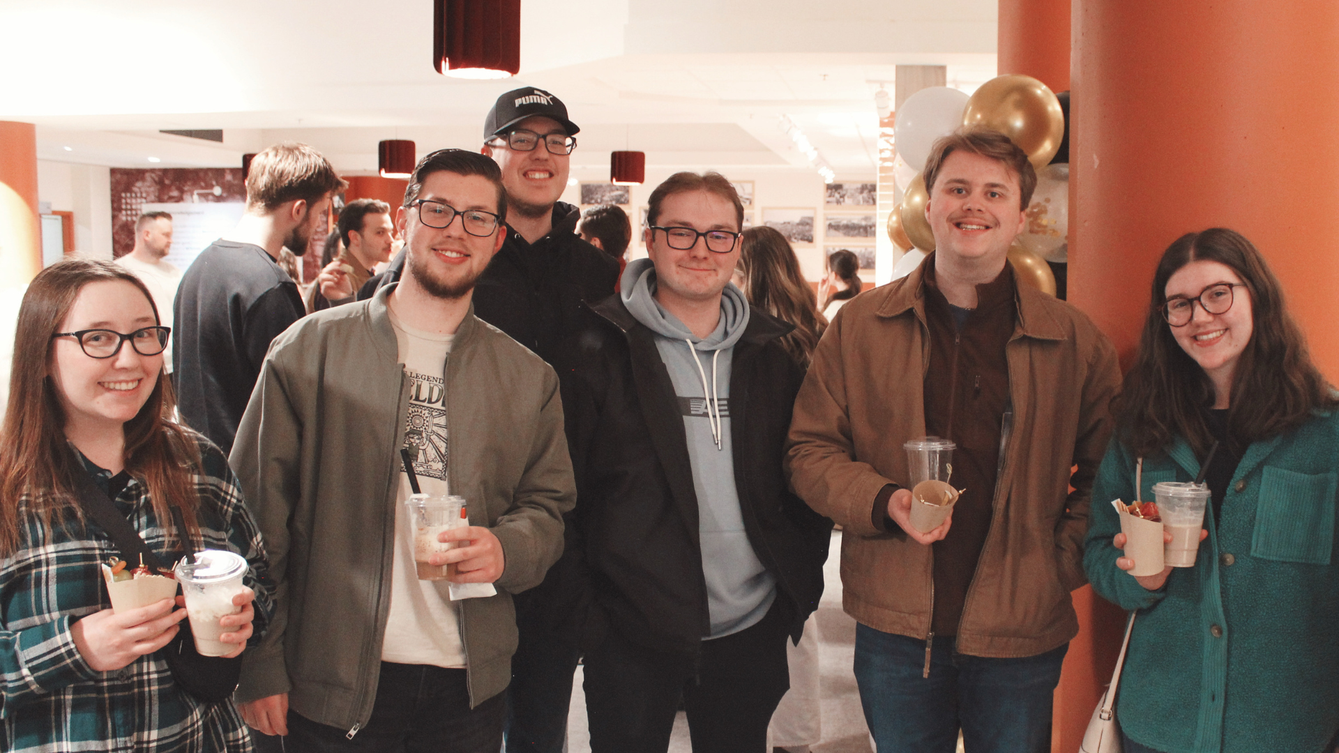 A group of six young adults smiling and standing together indoors, some holding drinks and snacks, with balloons and other people in the background.