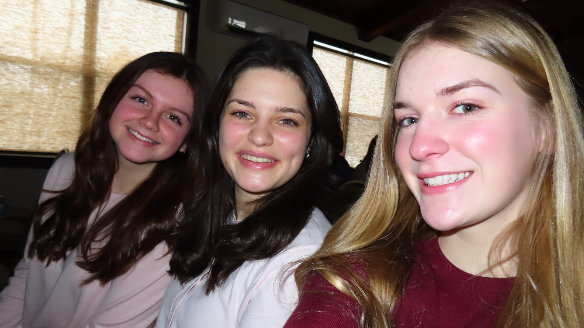Three young women taking a selfie together indoors, smiling, with windows in the background.