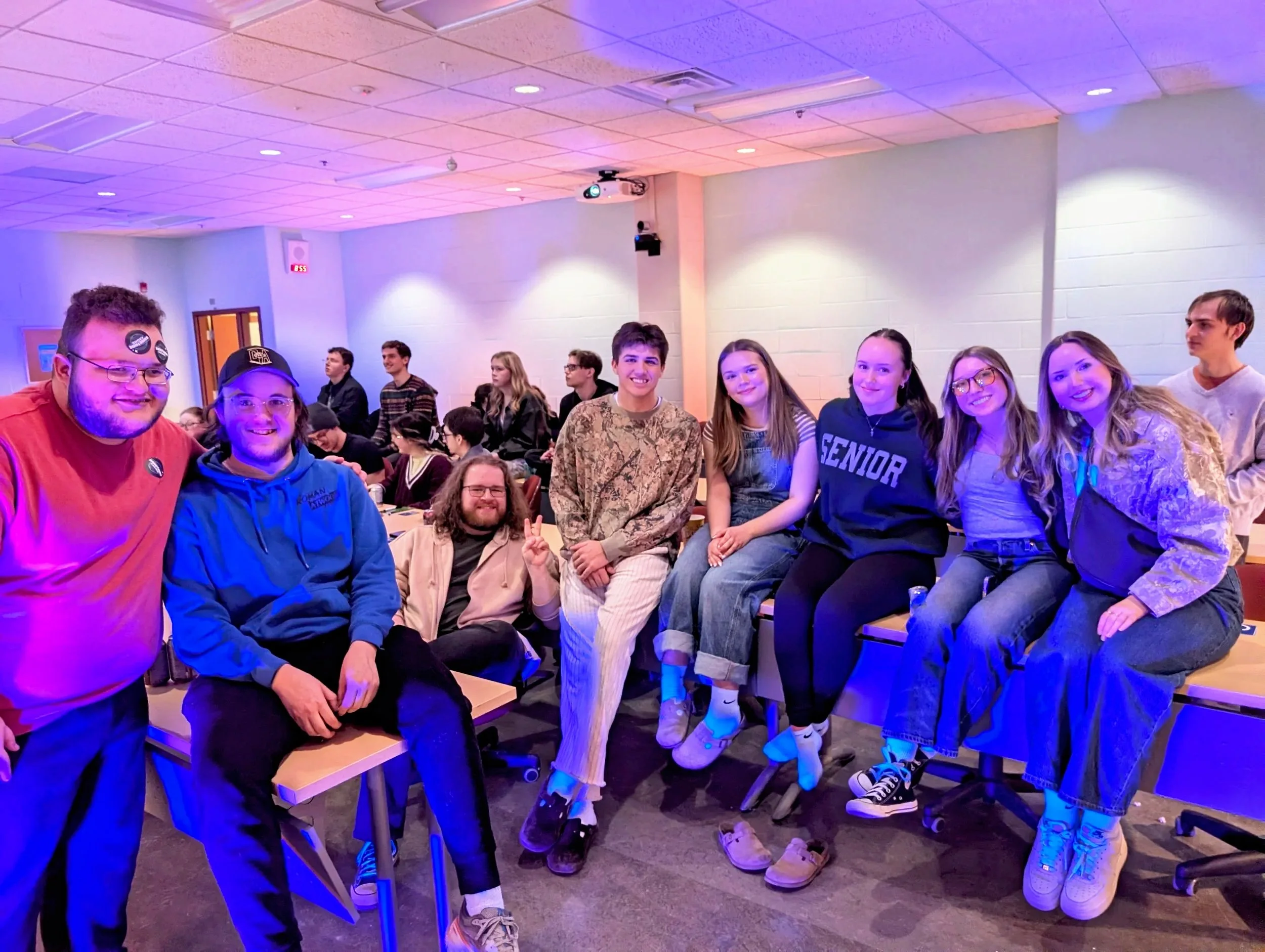 A group of young people gathered indoors, smiling and posing for the camera. Several are sitting at tables, some standing behind, with colorful lighting and a ceiling-mounted projector overhead.