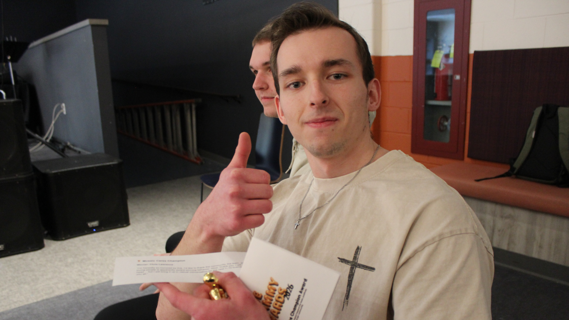 A young man with short brown hair giving a thumbs-up gesture, holding a piece of paper, wearing a beige t-shirt with a cross necklace, sitting in a room with a black speaker, a ladder, and a backpack in the background.