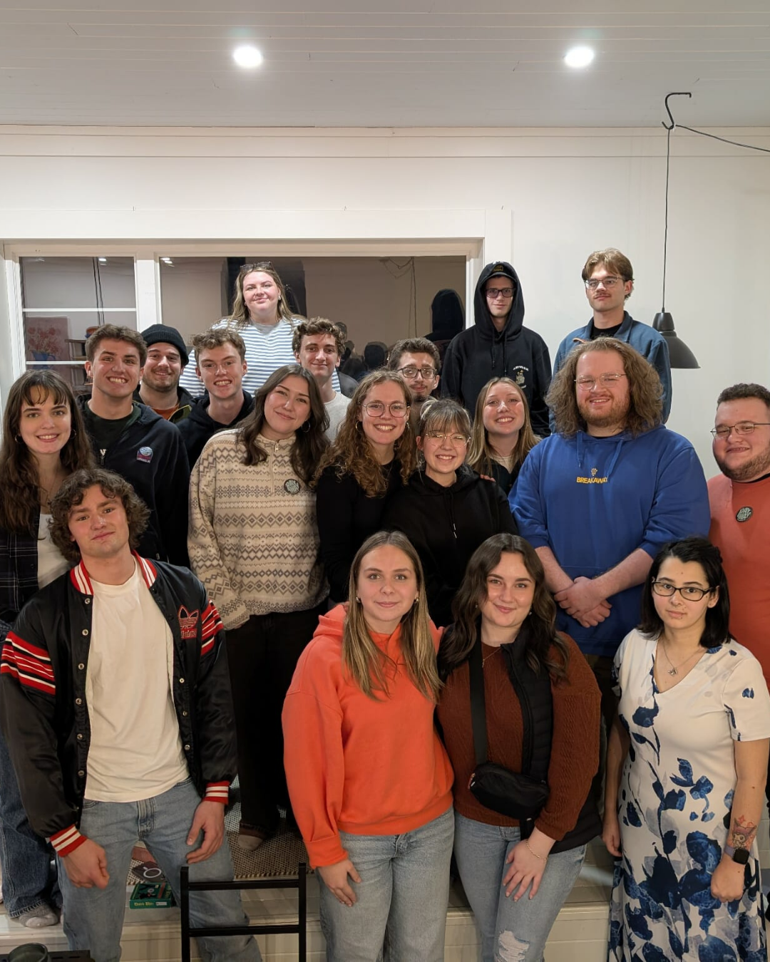 Group of 17 young adults gathered indoors, smiling for the photo, with some standing and others sitting, in a room with white walls and ceiling lights.