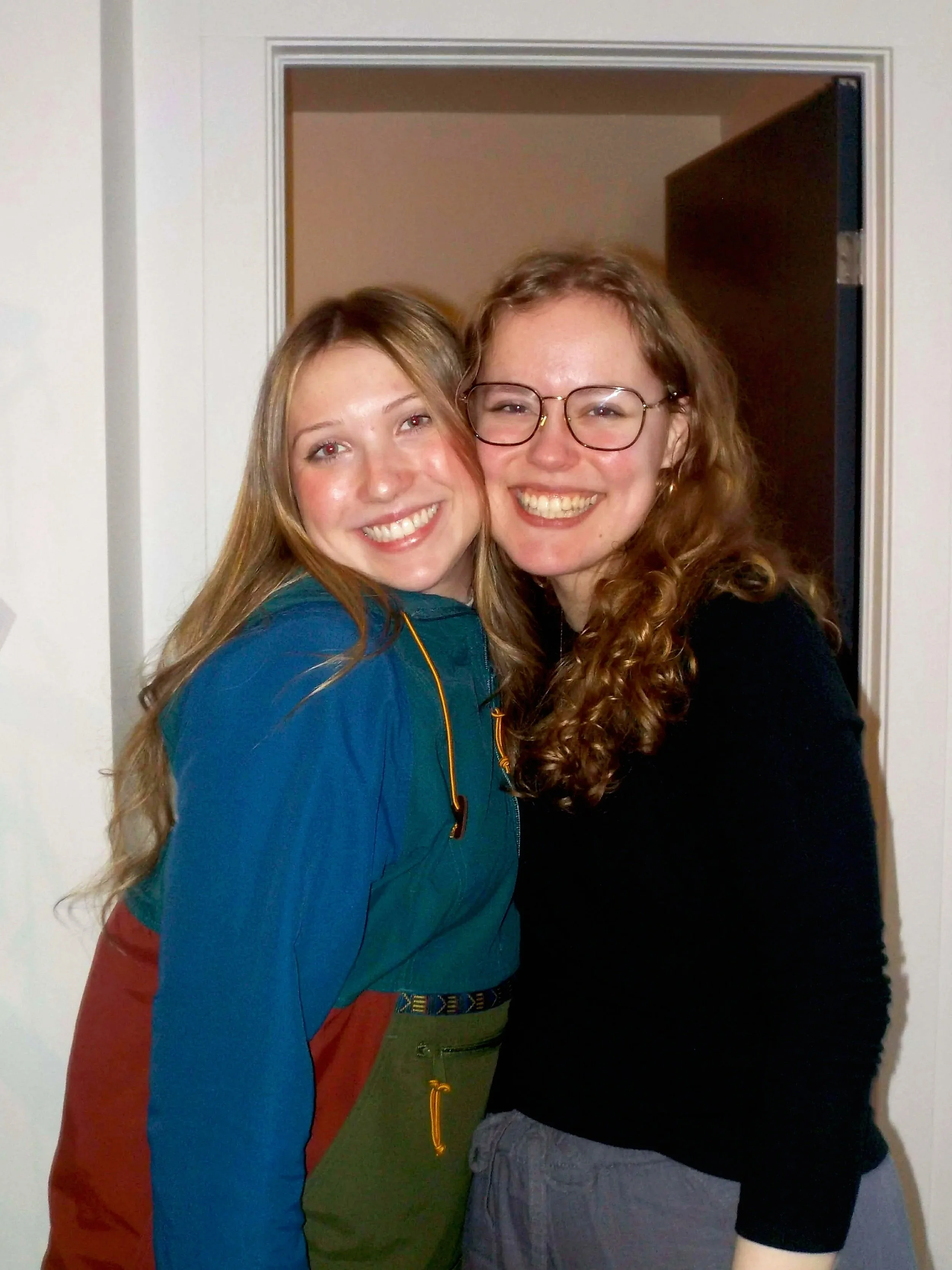 Two young women standing close together and smiling at the camera in front of an open doorway.