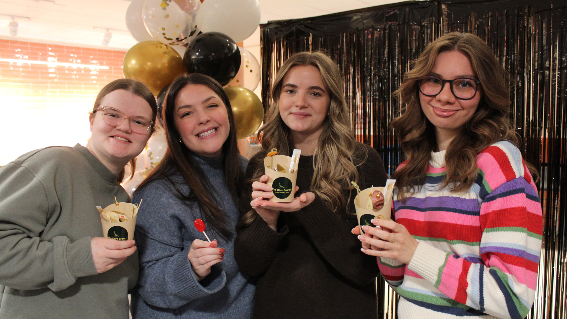 Four young women smiling and holding ice cream cups at a celebration, with balloons and a black and gold backdrop behind them.