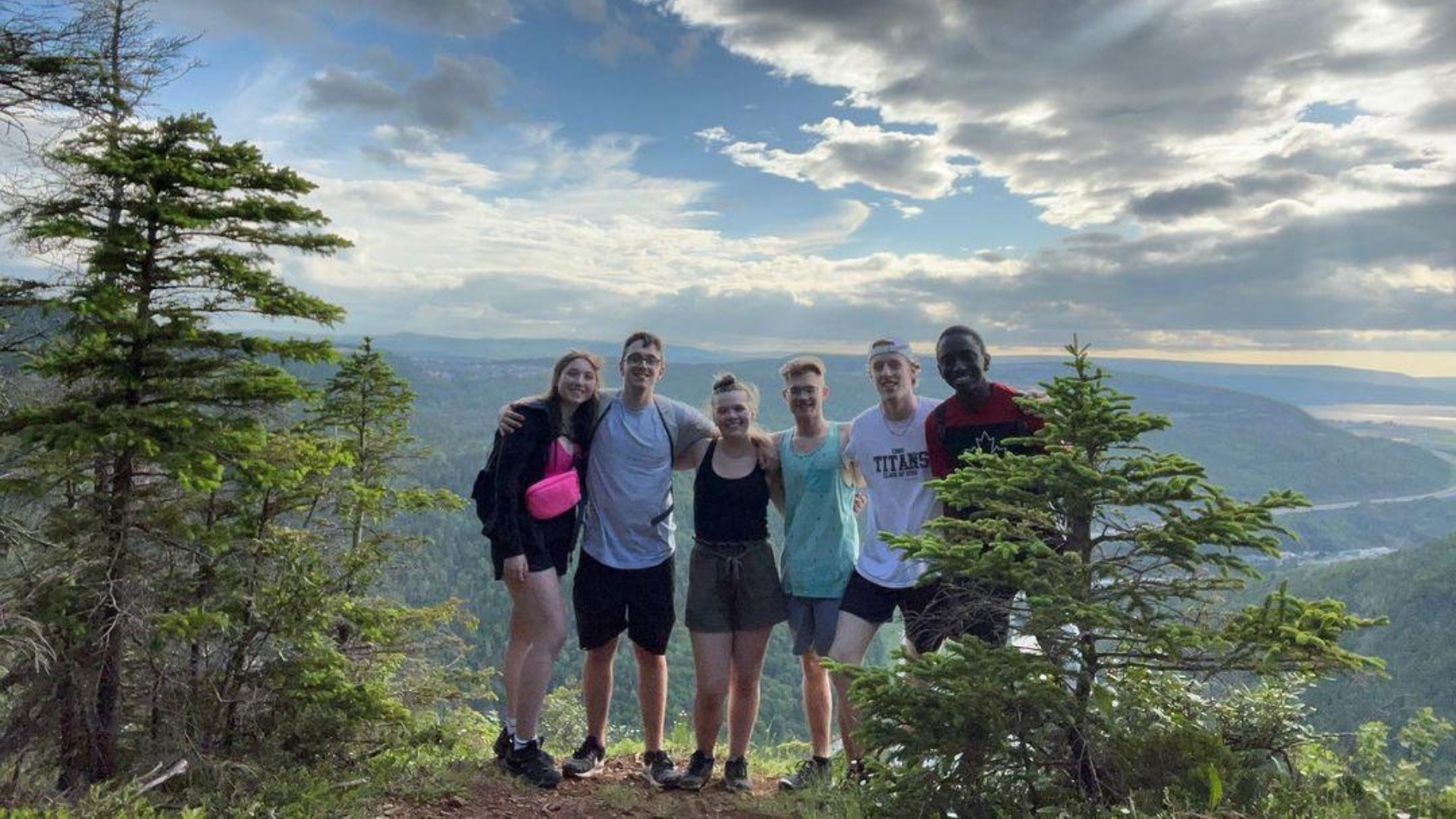 A group of seven young people standing on a mountain trail with trees and a scenic landscape of hills, water, and cloudy sky in the background.