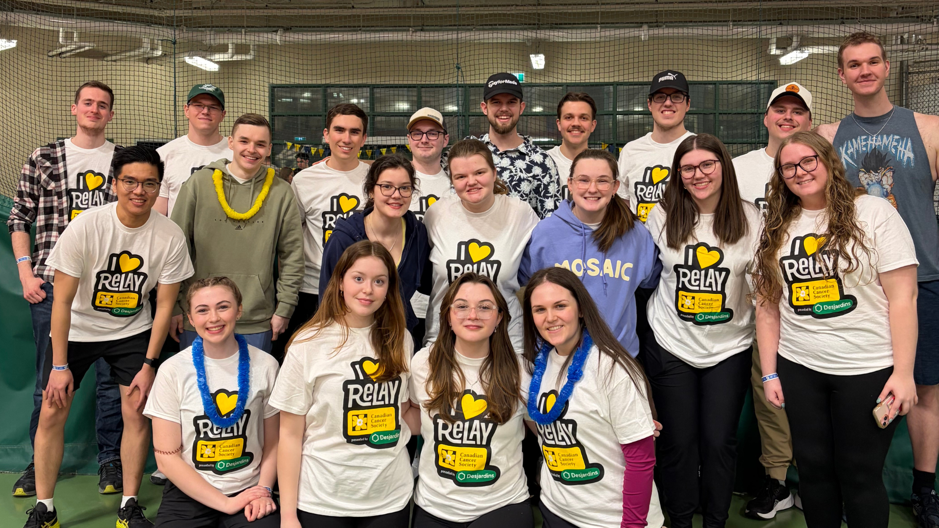 A group of young people at an indoor sports event, wearing Relay for Life shirts and leis, standing in front of a net with smiles.
