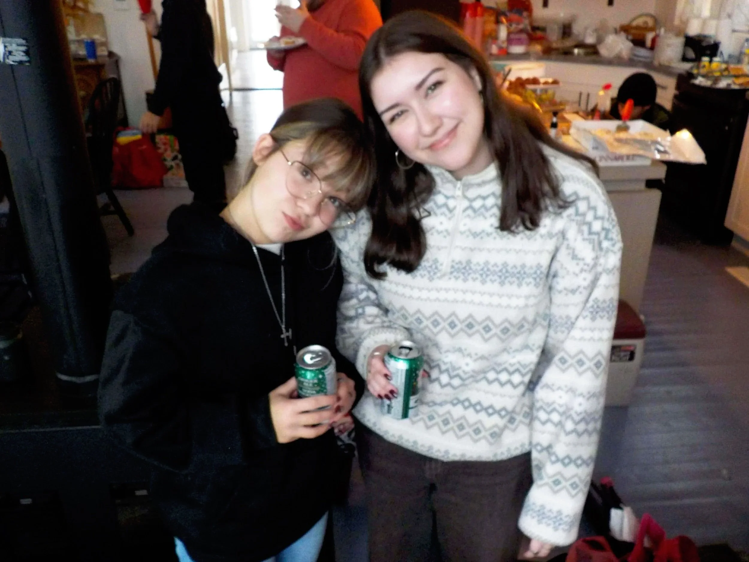 Two women holding cans, one with glasses and a black top, the other with earrings and a patterned sweater, at a casual indoor gathering with a busy kitchen in the background.