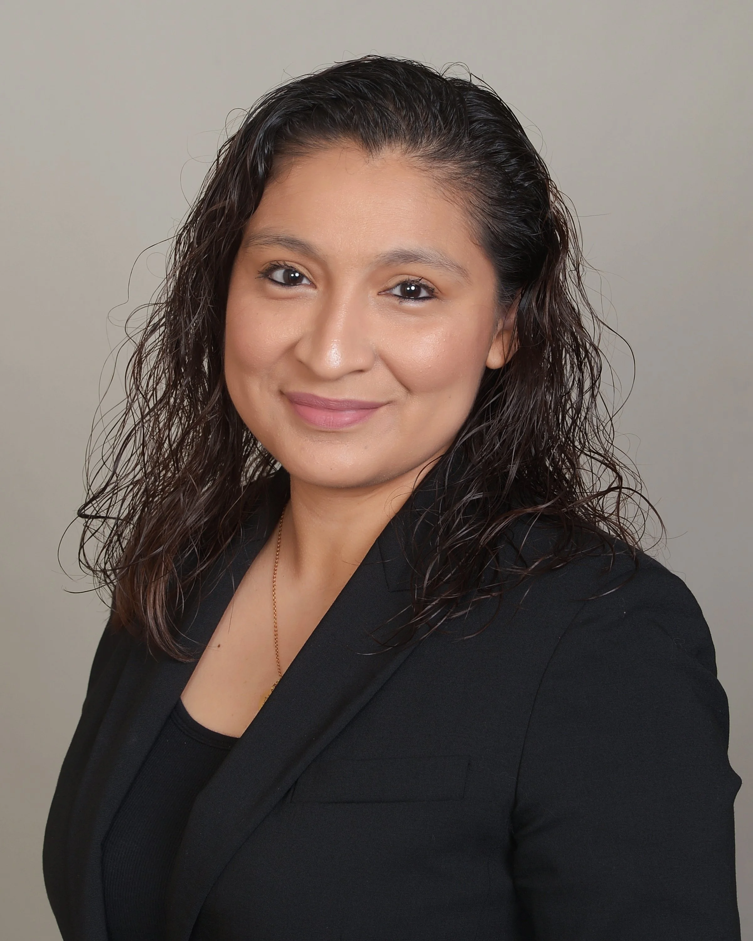Close-up of a woman with dark, wavy hair, wearing a black blazer and a necklace, smiling at the camera against a plain background.