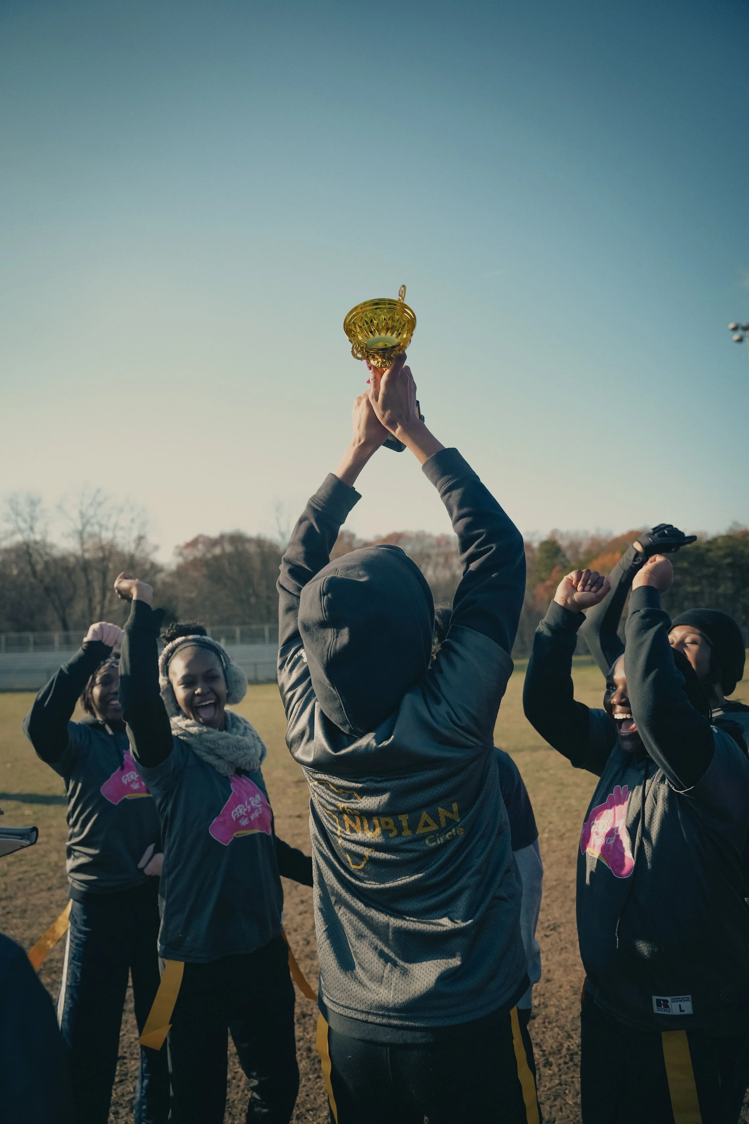 A group of young women in athletic wear celebrating outdoors after a race, holding a trophy up in the air, on a field with trees in the background on a clear day.