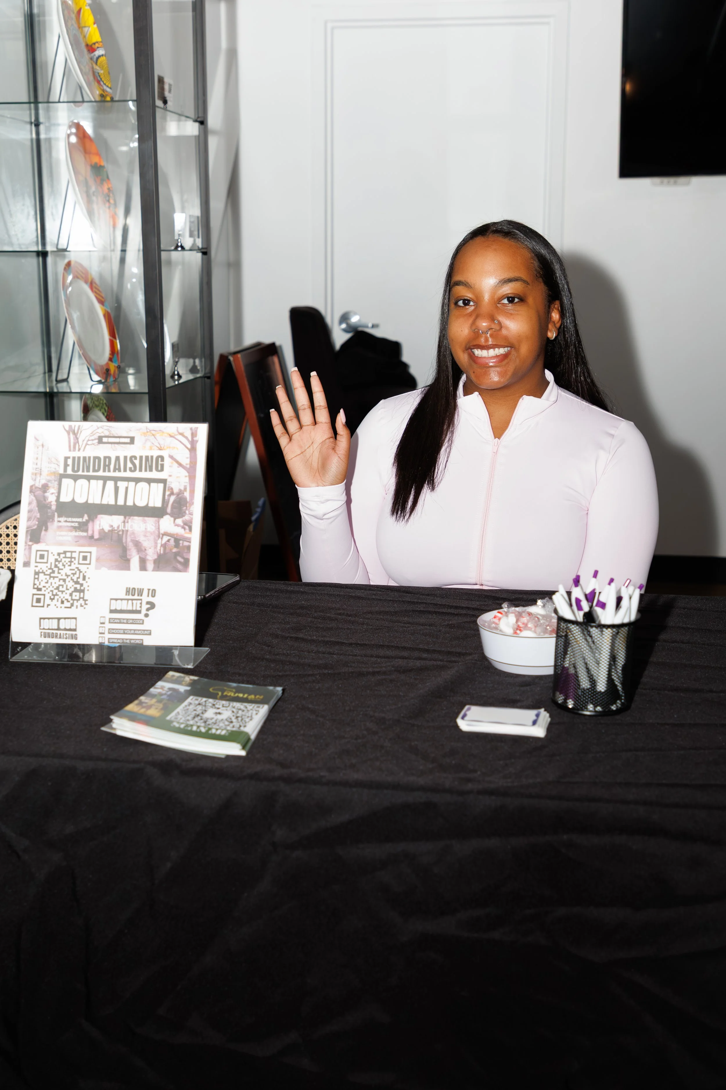 A woman with long black hair and a nose piercing, wearing a white zip-up jacket, is smiling and waving at a fundraising event. There is a table in front of her with promotional materials, a bowl of candy, and a container holding pens. Behind her, a glass display case with decorative plates and a black chair can be seen.