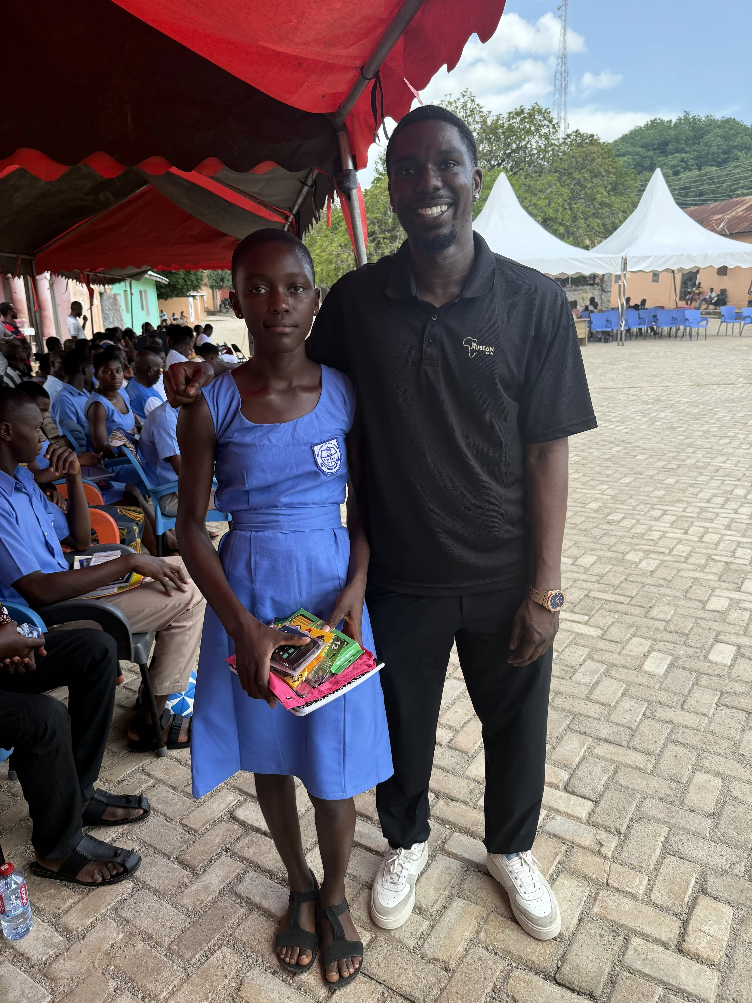 A young girl in a blue school uniform stands next to a smiling man in black clothing at an outdoor event. The girl holds a tray with school supplies, and there are other children and adults seated in the background under tents.