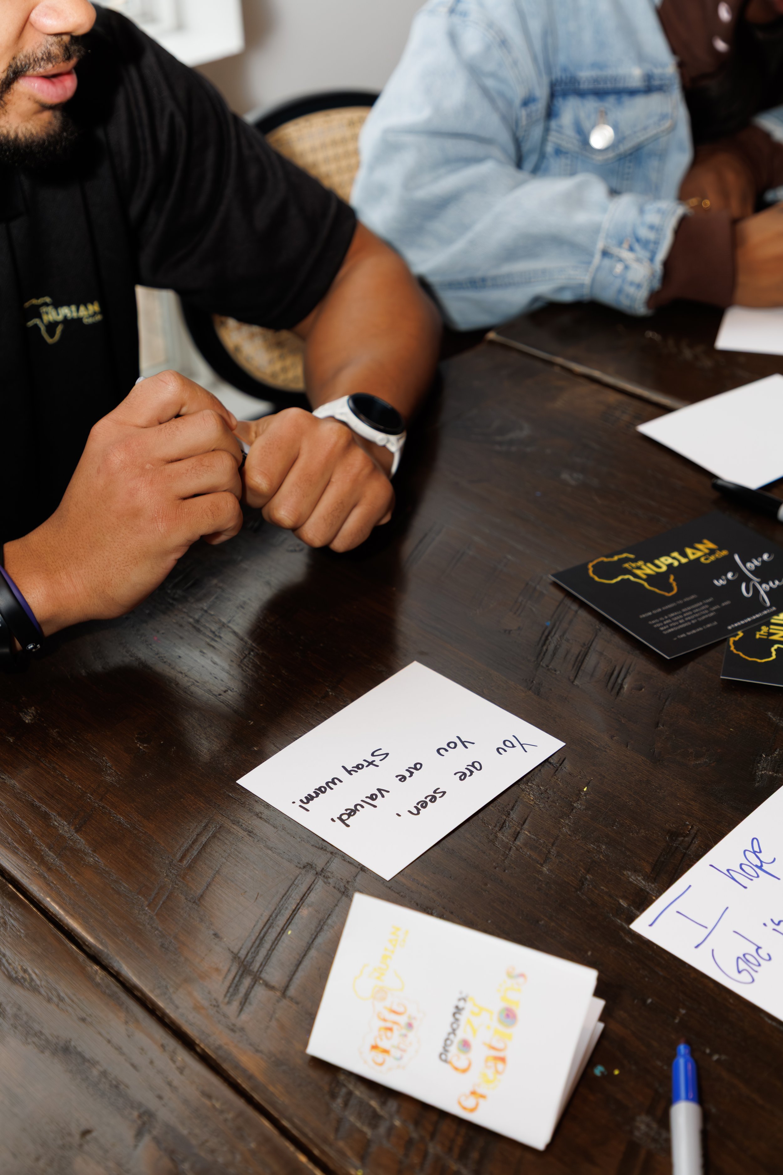 Close-up of a wooden table with handwritten notes and drawings, and two partially visible people are sitting at the table. One person is wearing a black shirt and a smartwatch, and the other is wearing a denim jacket. The notes contain colorfully written messages and slogans supporting Nigeria, with one visible note reading: "We sent, you are valued. Say Well!"
