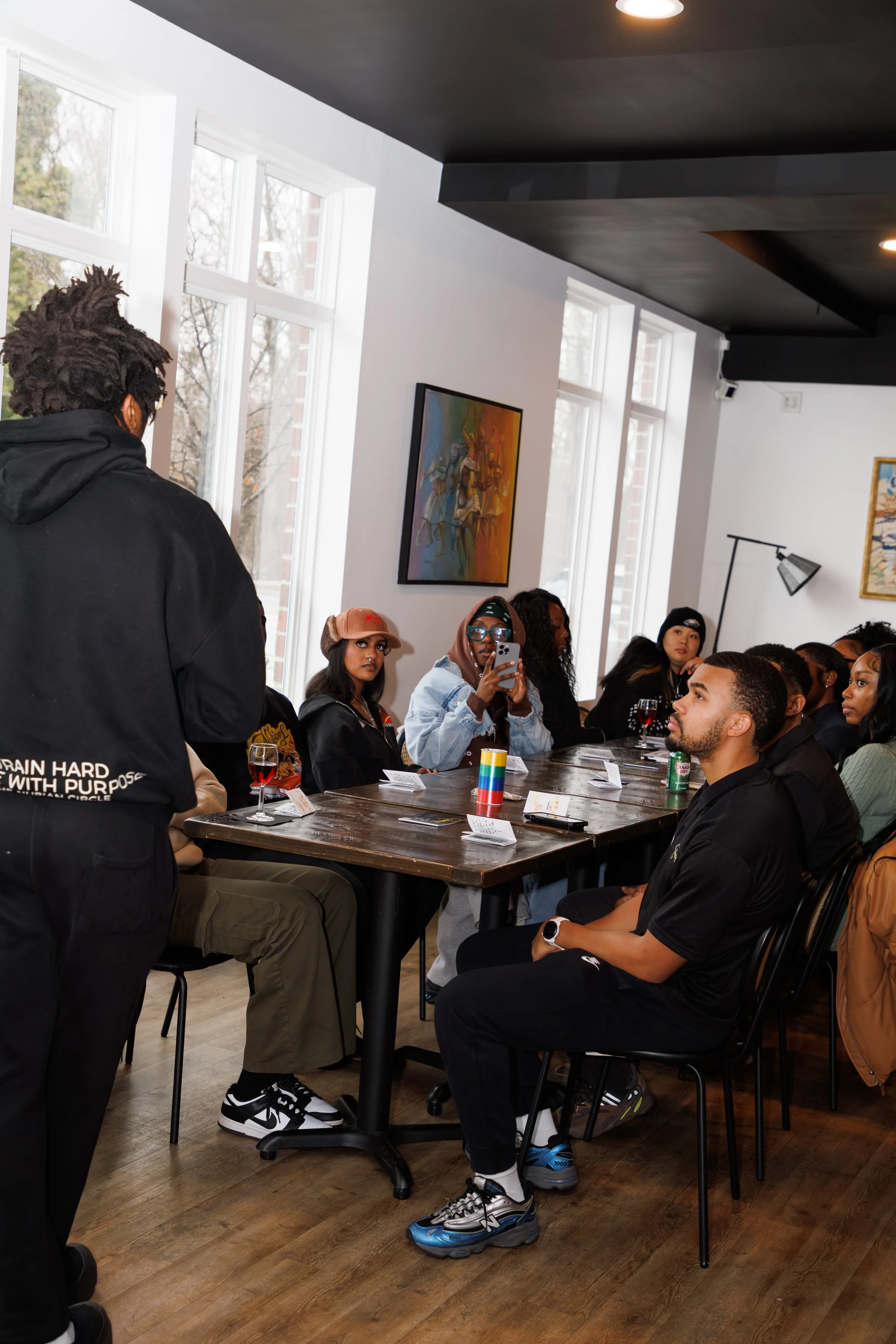 People sitting and listening in a restaurant or cafe, some taking notes or photos, with a man standing and speaking to the group, decorators on the walls, large windows, wooden floor.
