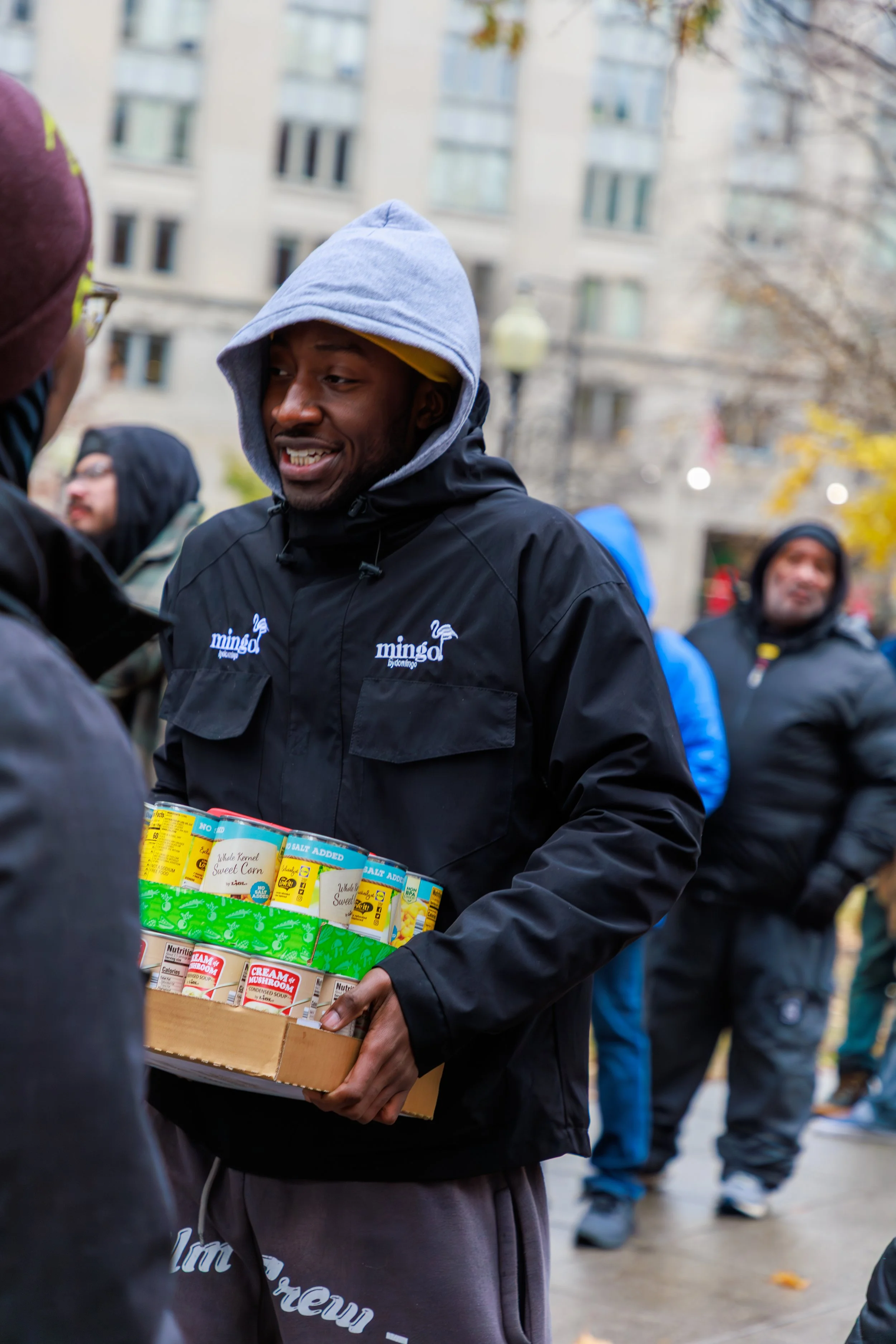 A man wearing a gray hoodie and black jacket is holding a box of canned goods, including corn, salt, mushroom soup, and other items. He appears to be in a public outdoor space with other people present.