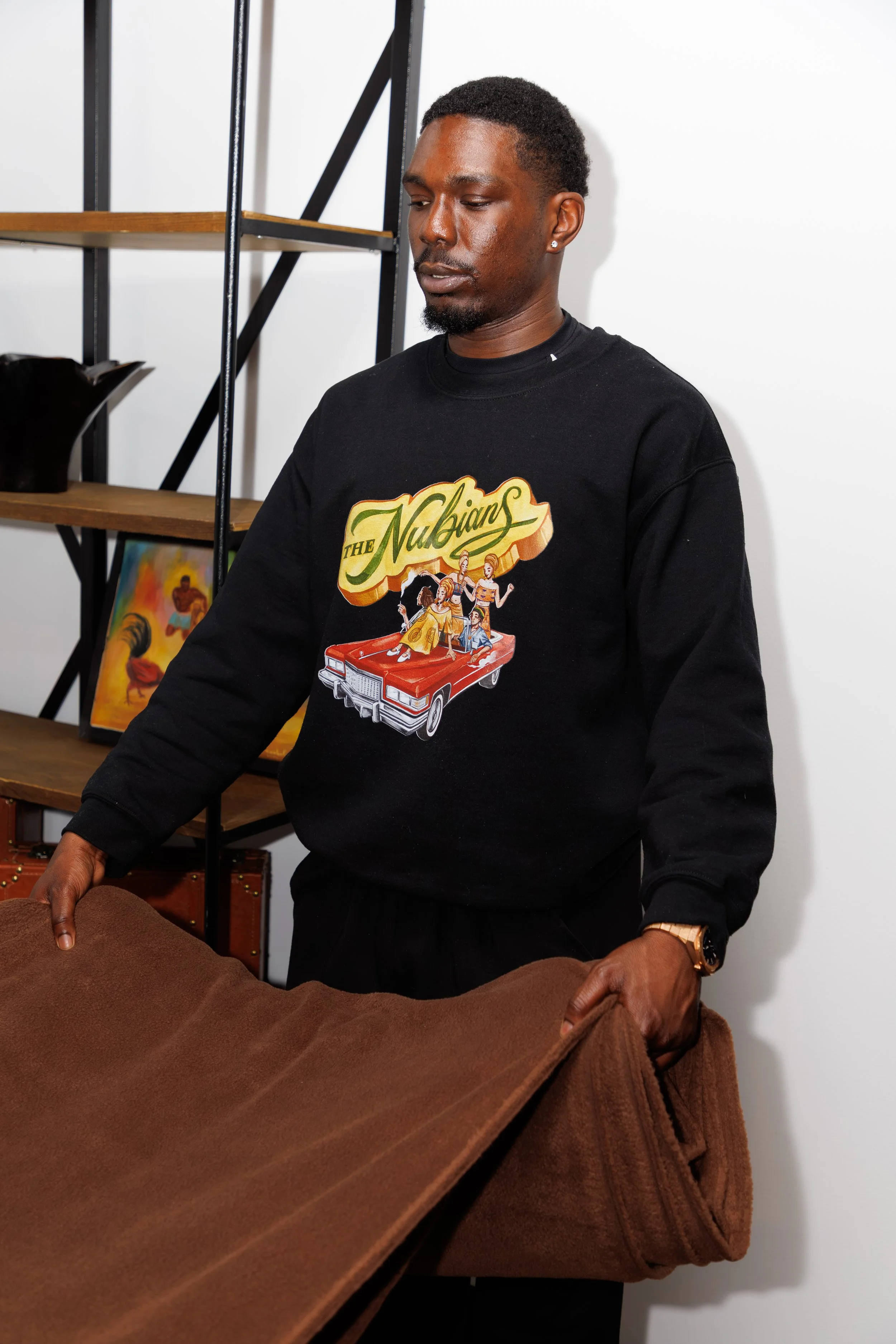 A man with dark skin, black hair, and a beard wearing a black sweatshirt with a graphic design of women and a car, standing next to a brown piece of cloth.