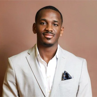 Portrait of a young man in a light gray suit and white shirt, standing against a plain brown background.