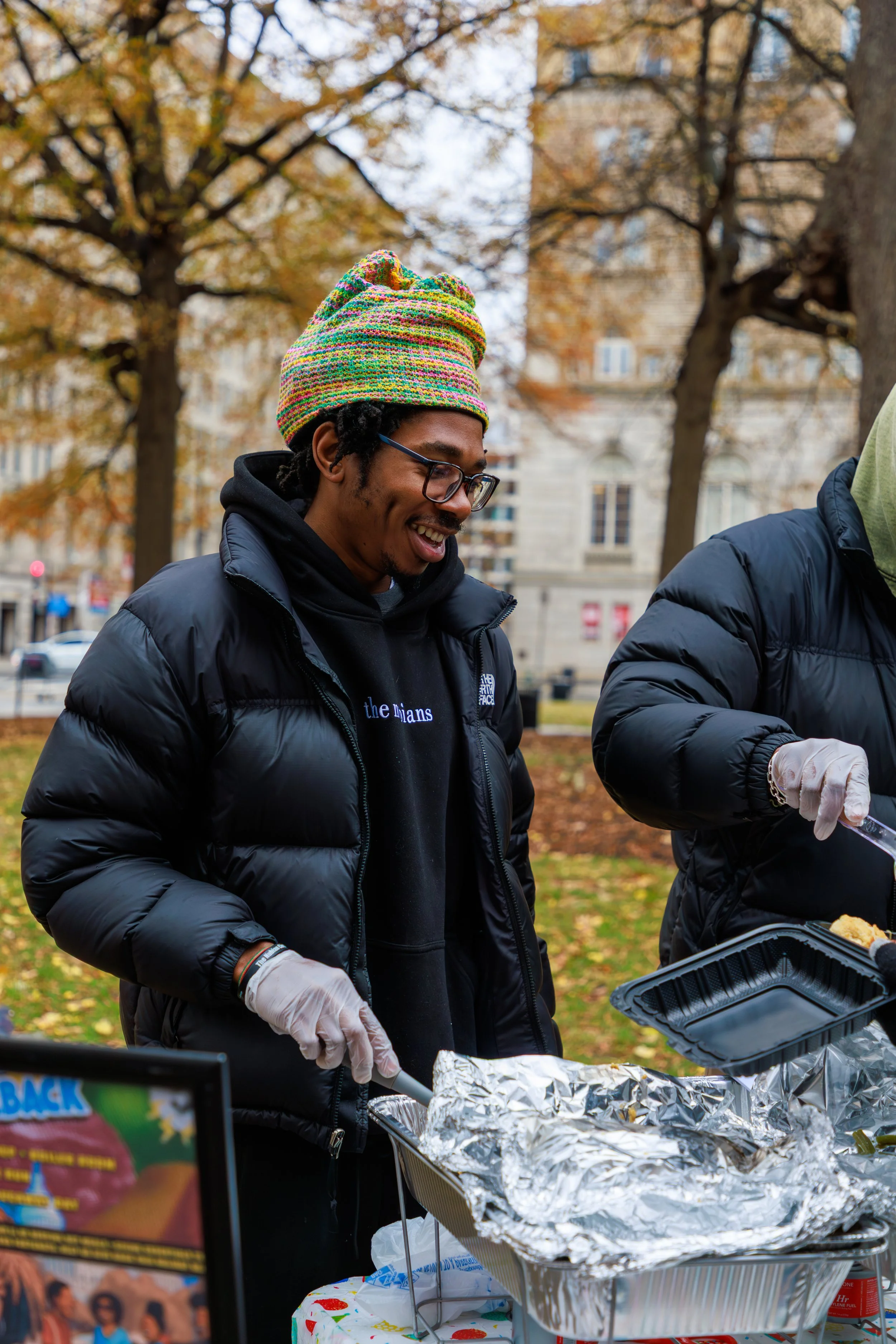 A young man with glasses and a colorful knitted beanie is smiling while serving food at a barbecue in a park. He is wearing a black jacket and gloves, and there are aluminum foil-covered trays of food in front of him. Other people and trees with fall foliage are visible in the background.