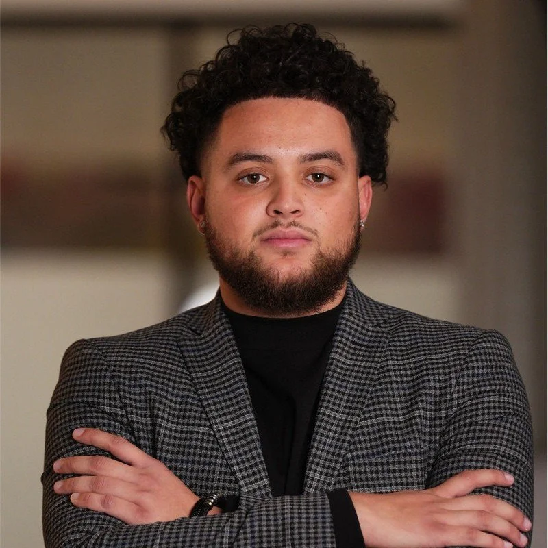 A young man with curly hair and a beard, wearing a gray checkered blazer and black shirt, standing indoors with arms crossed and a neutral expression.