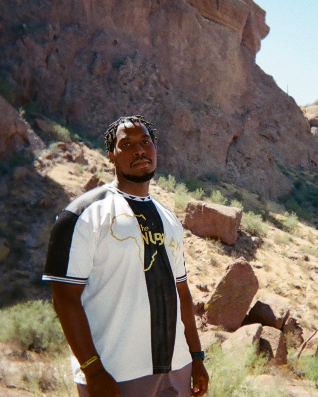 A man with braids and a goatee standing outdoors in a desert landscape with rocks and sparse vegetation, wearing a white and black jacket and a black T-shirt.