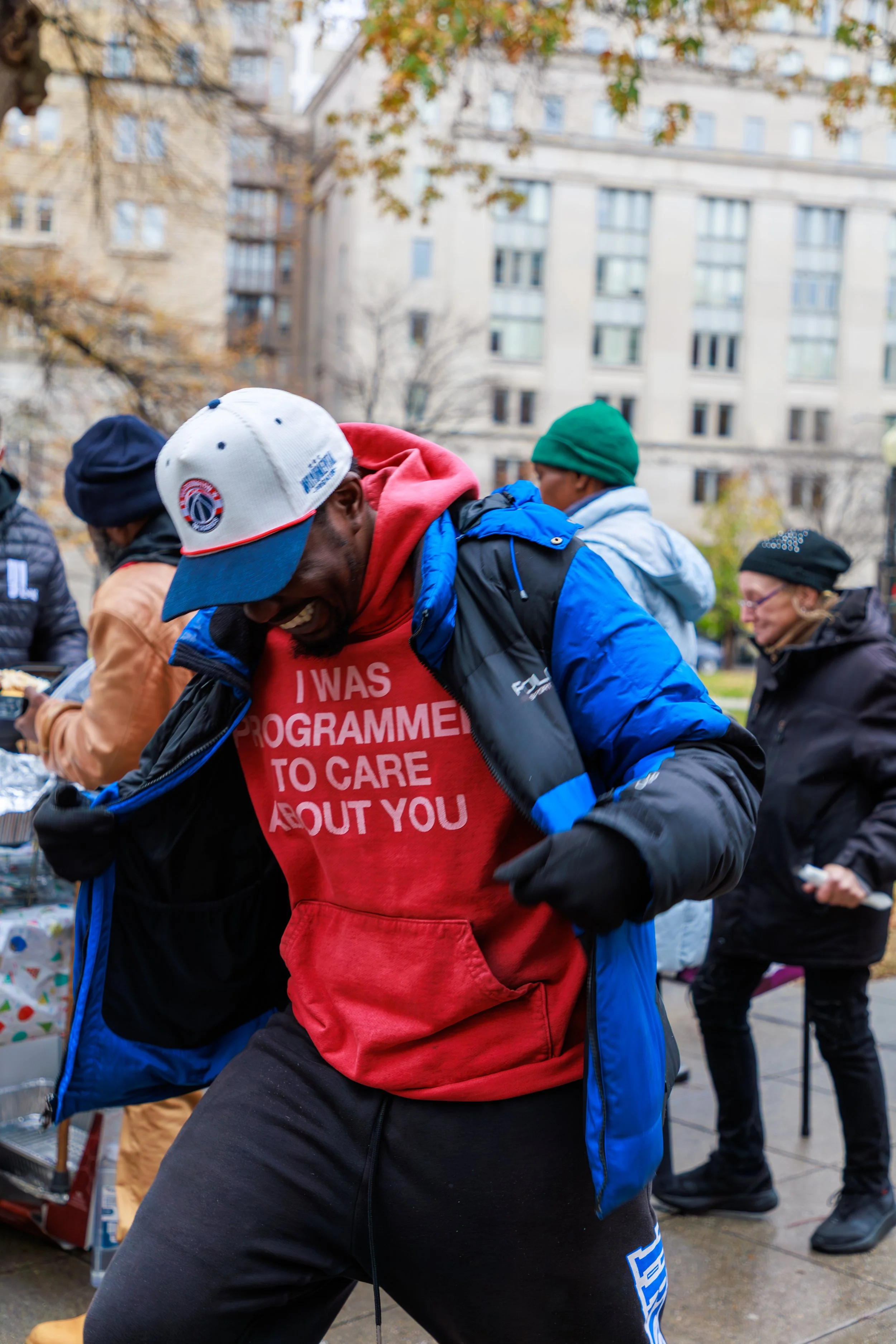 People participating in an outdoor event, wearing jackets and hats, with a man in the foreground wearing a red hoodie that says 'I was programmed to care about you.' in an urban setting with trees and buildings in the background.