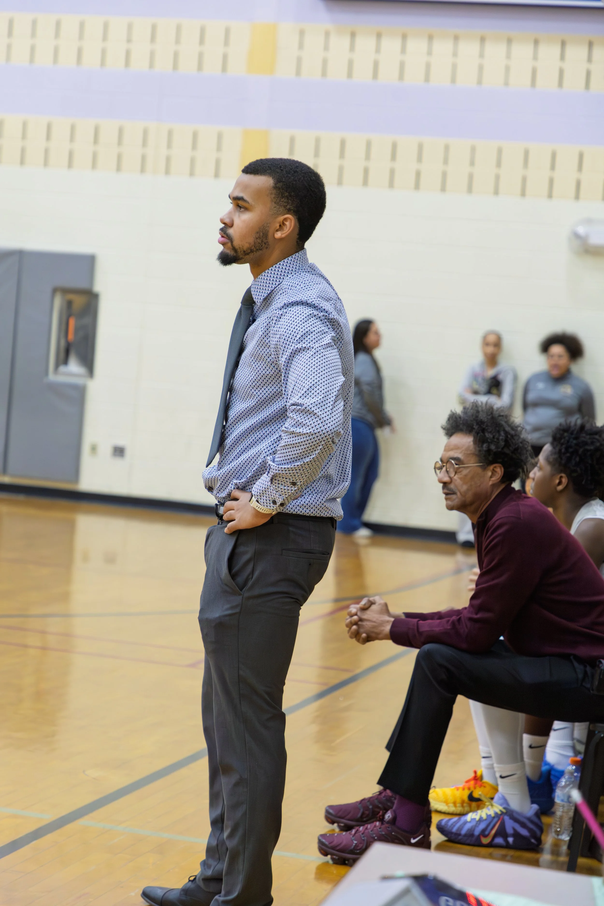 A man standing in a gymnasium, praying with his hands clasped, while people and athletes are seated and watching in the background.