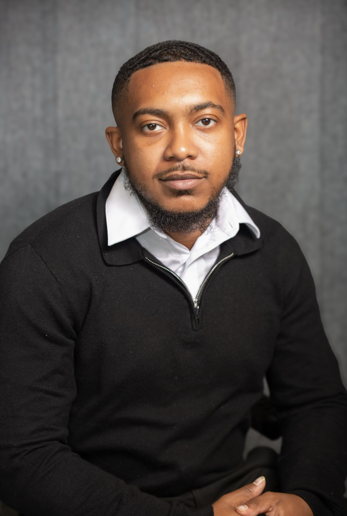 A young man with a short haircut and a beard, wearing a white collared shirt under a black zip-up sweater, sitting against a gray background.
