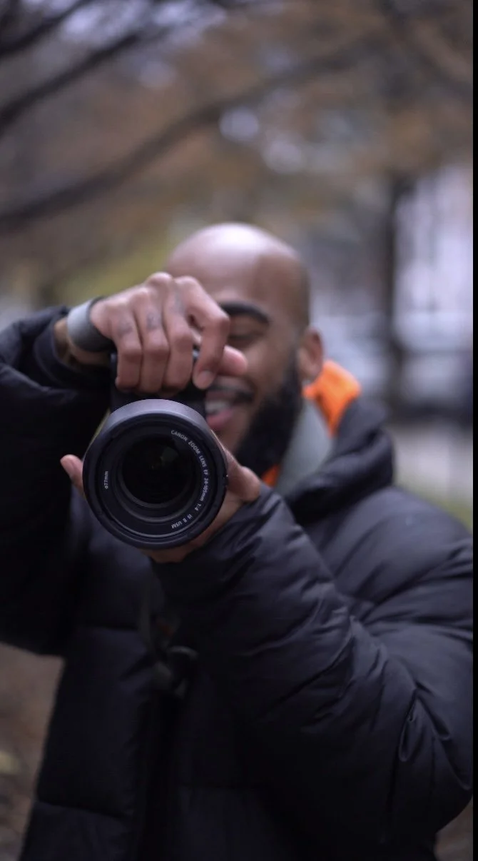 Man taking a selfie outdoors with a professional camera, smiling, with trees in the background.
