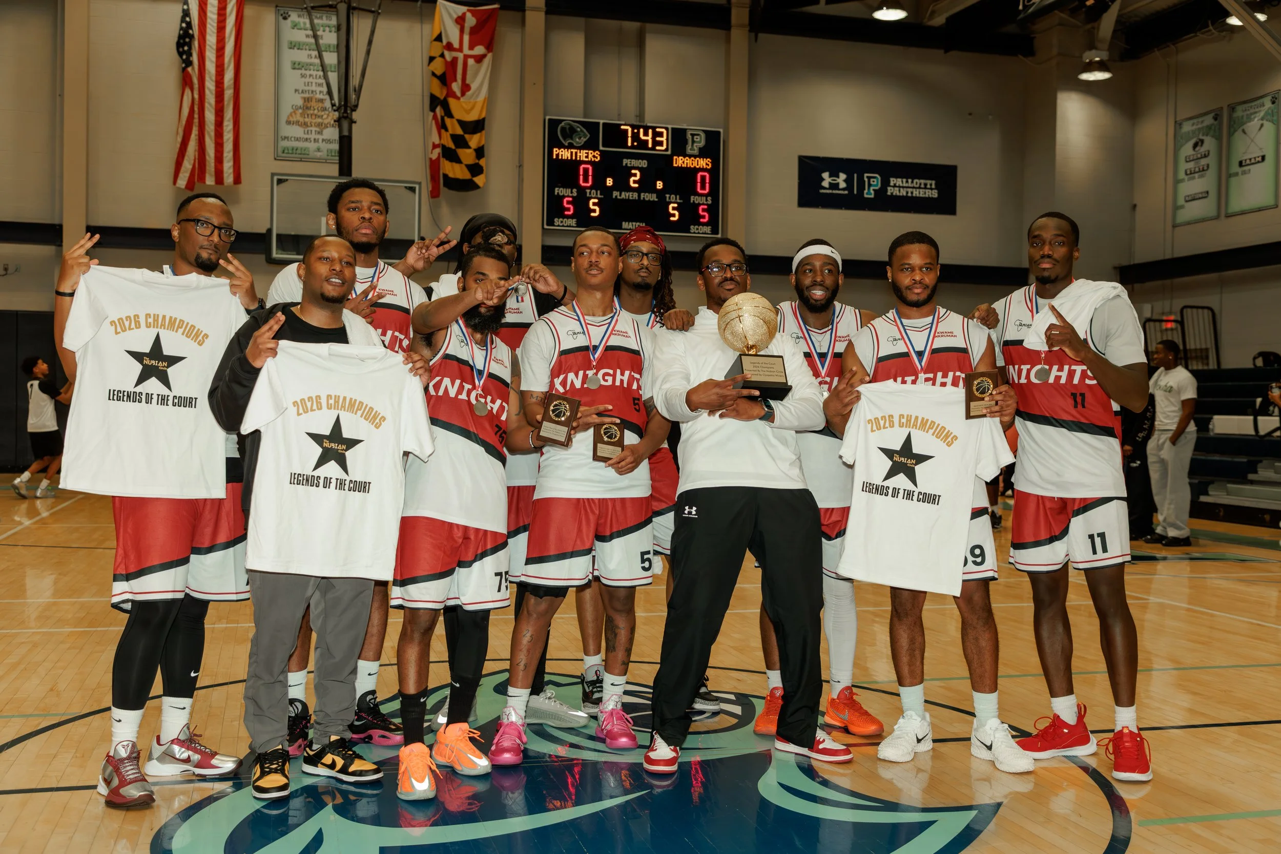 A basketball team celebrating a victory on the court, holding trophies and wearing medals. The scoreboard shows they won with a score of 2-0. The players are dressed in team uniforms, some holding shirts that read '2026 Champions Legends of the Court,' and some making victory signs. The gym is decorated with flags and banners.