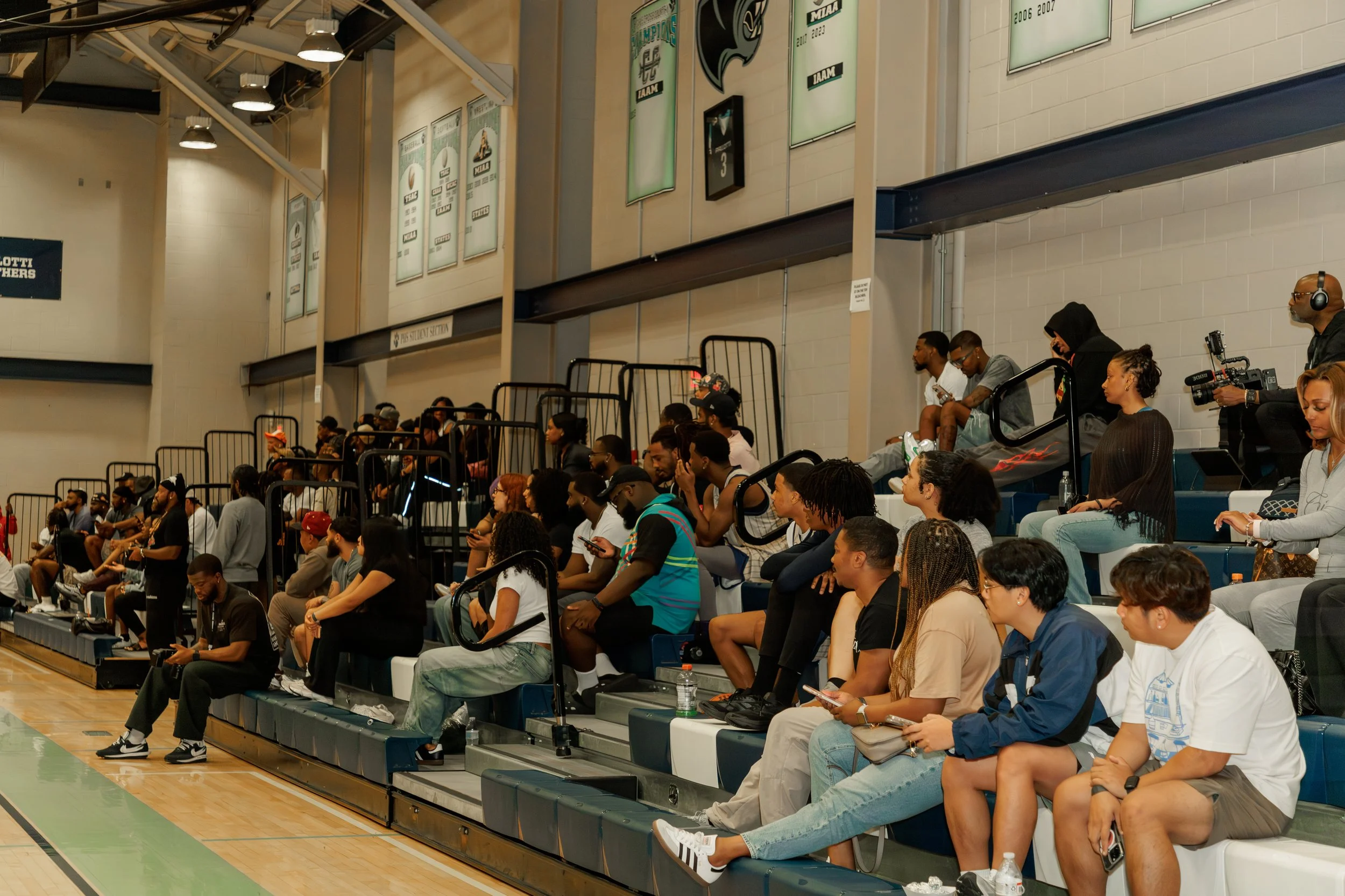 A group of spectators sitting and standing in the stands of a sports arena, watching an event on the court. Some are using their phones, and a camera operator wearing headphones is filming.