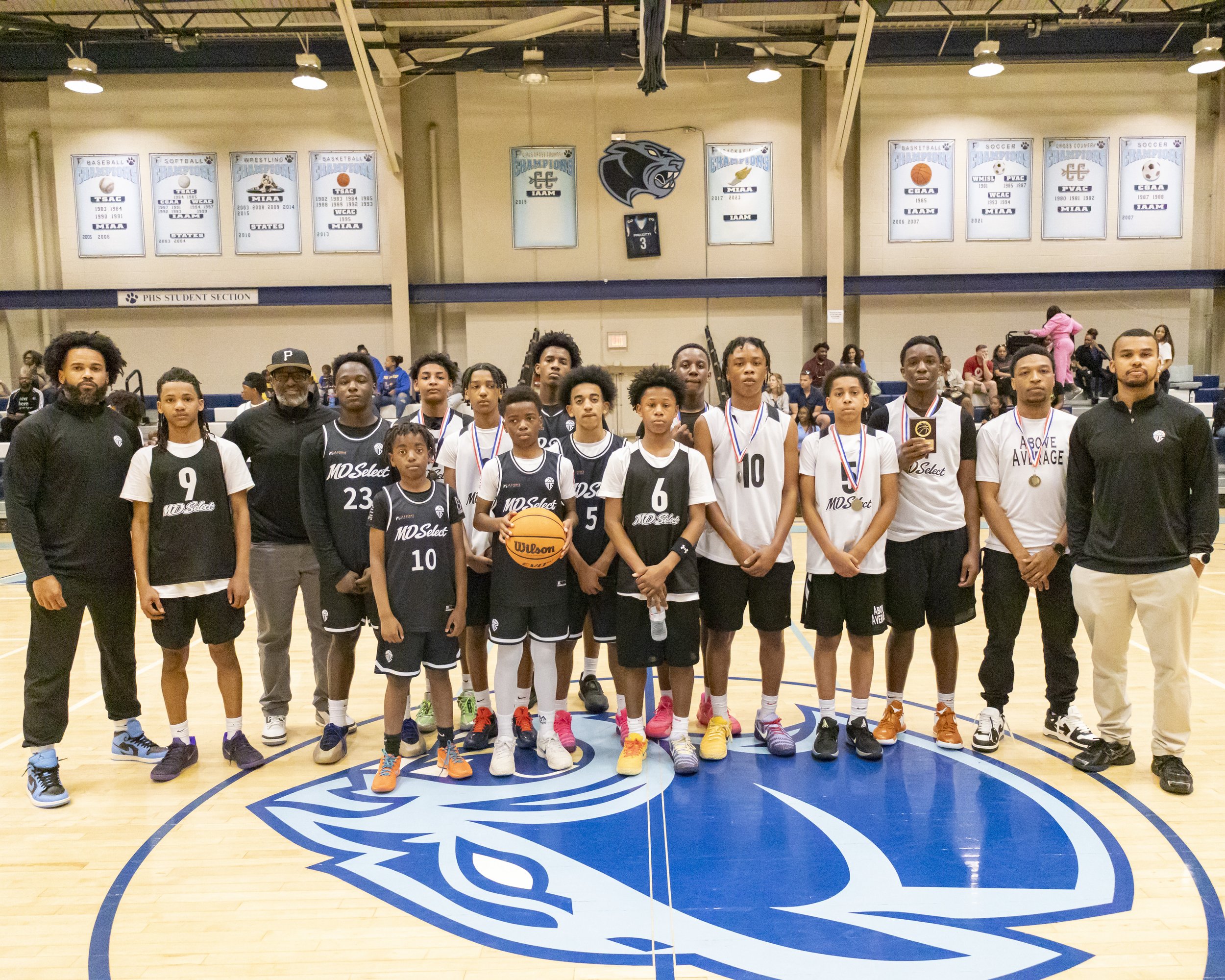 A group of young basketball players and coaches posing for a photo on an indoor court, with banners and team logos displayed on the wall behind them.