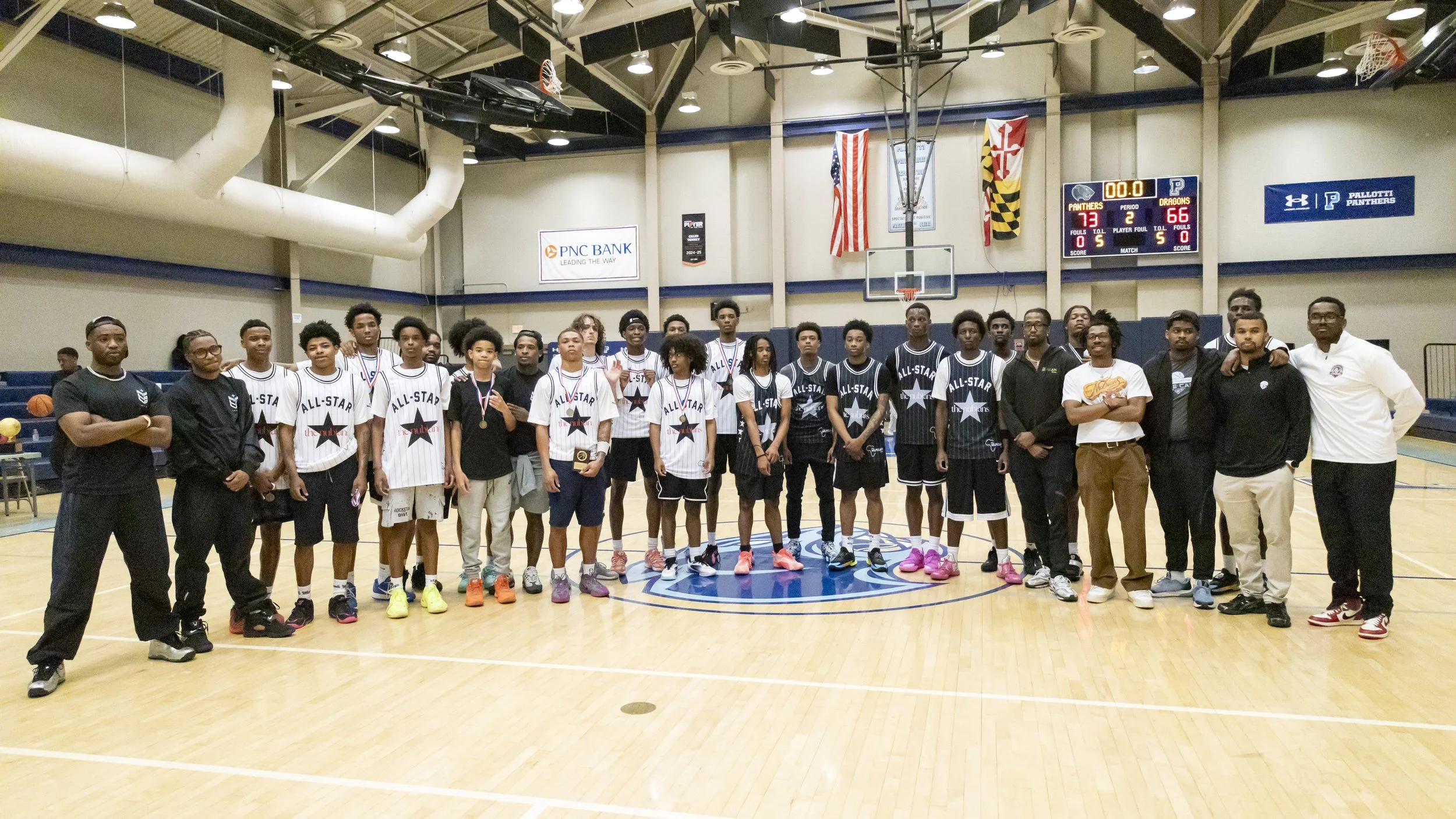 A group of basketball players and coaches standing on a basketball court inside a gymnasium, with scoreboard displaying 73-66 and the game clock at 00.0, celebrating after a game.
