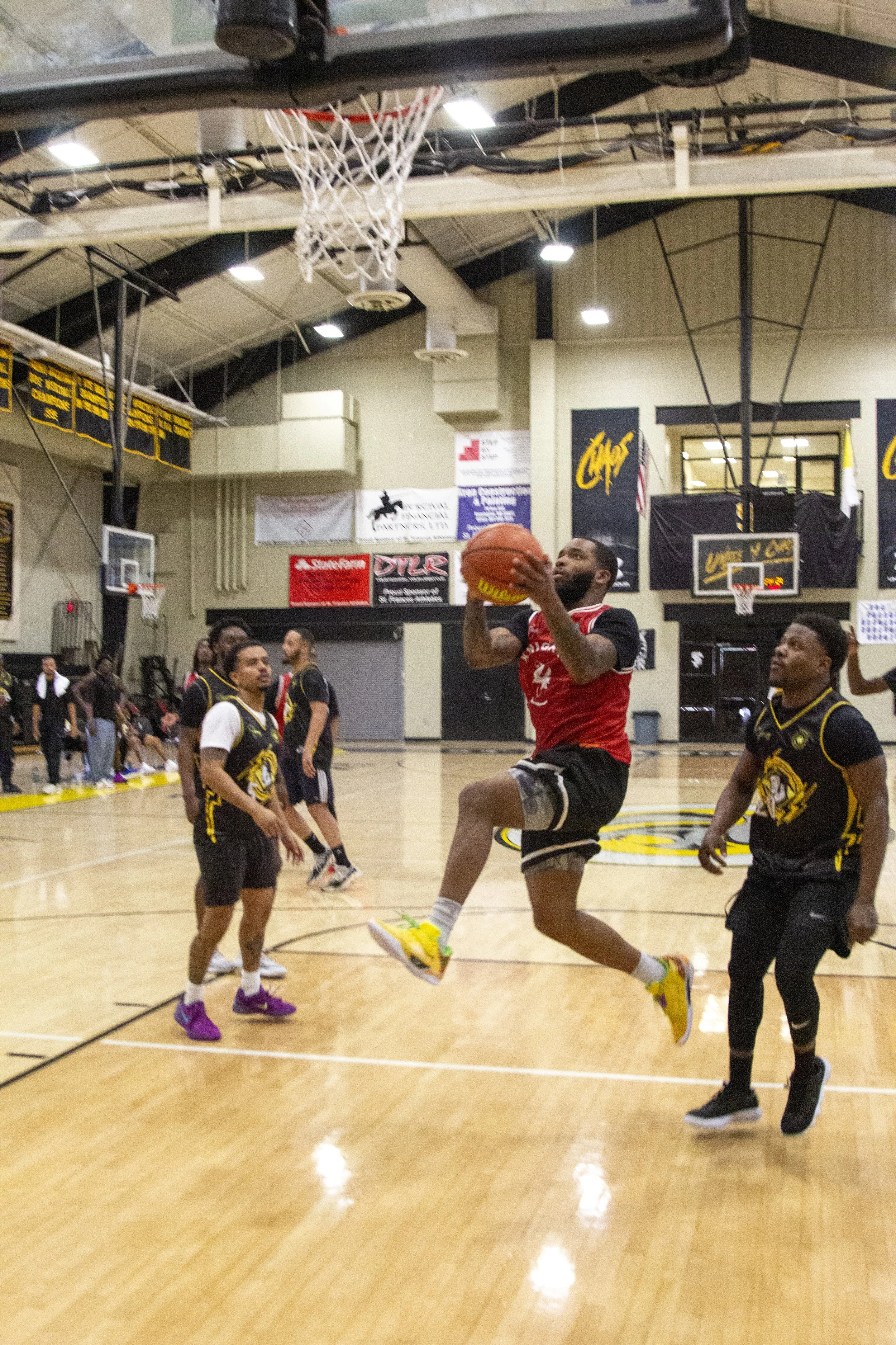 A man in a red basketball jersey and black shorts is jumping to make a shot in an indoor basketball court. Several players around him are watching or preparing. The court has banners on the walls and a basketball hoop overhead.