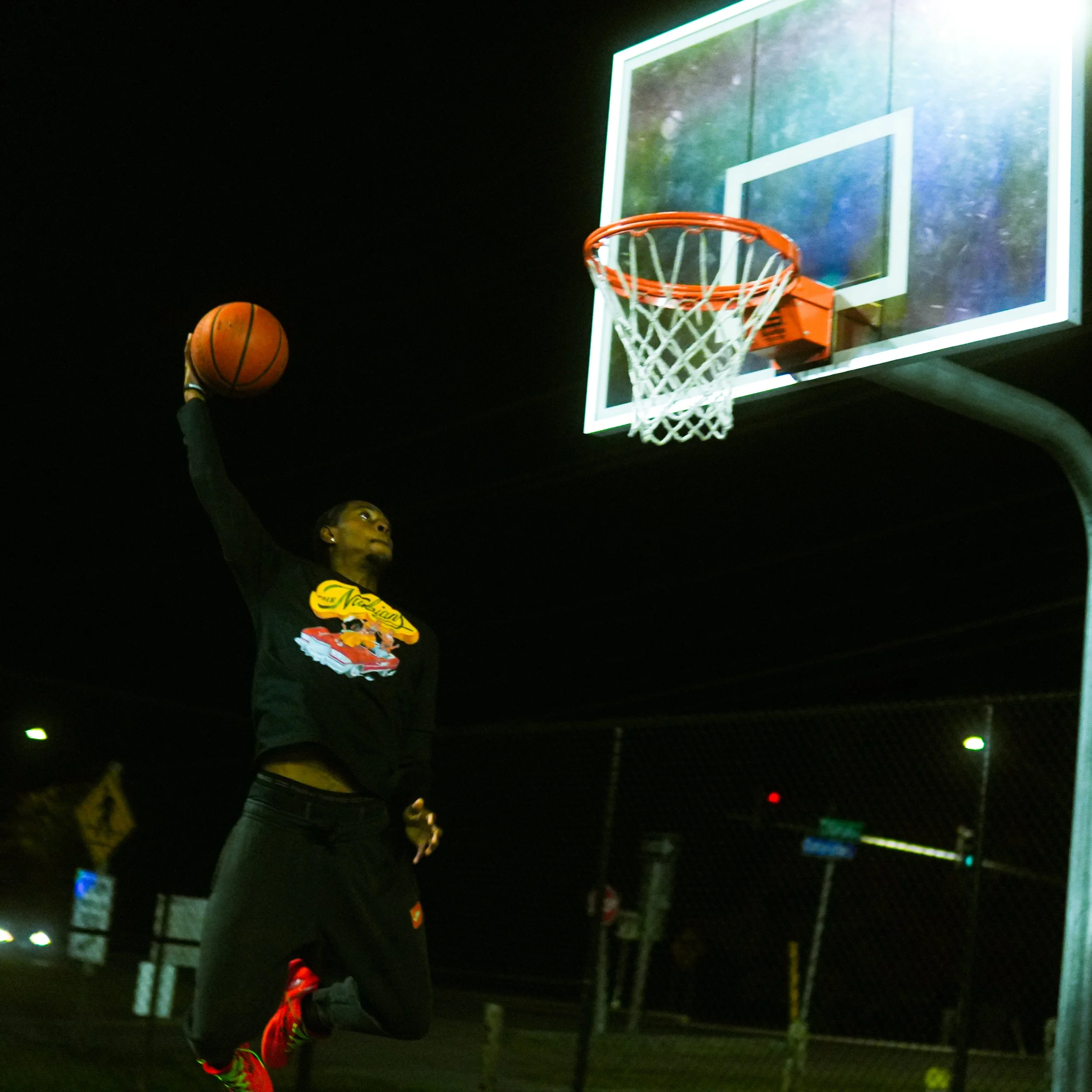 Basketball player taking a shot at night under a lit basketball hoop with a backboard.