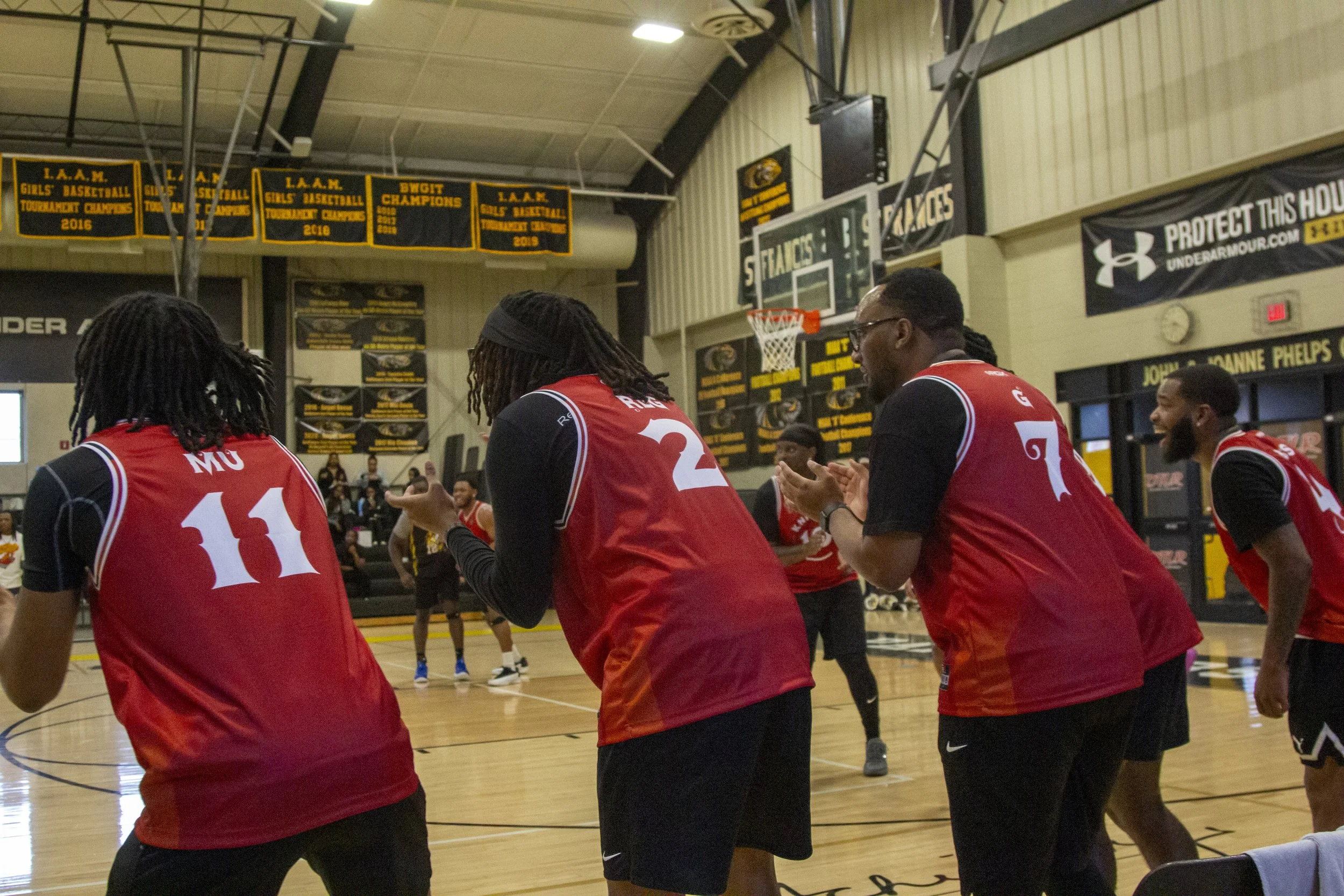 A group of female basketball players wearing red jerseys with white numbers are standing in a gymnasium, appearing to celebrate or pray during a game or practice. There are banners and a scoreboard in the background.