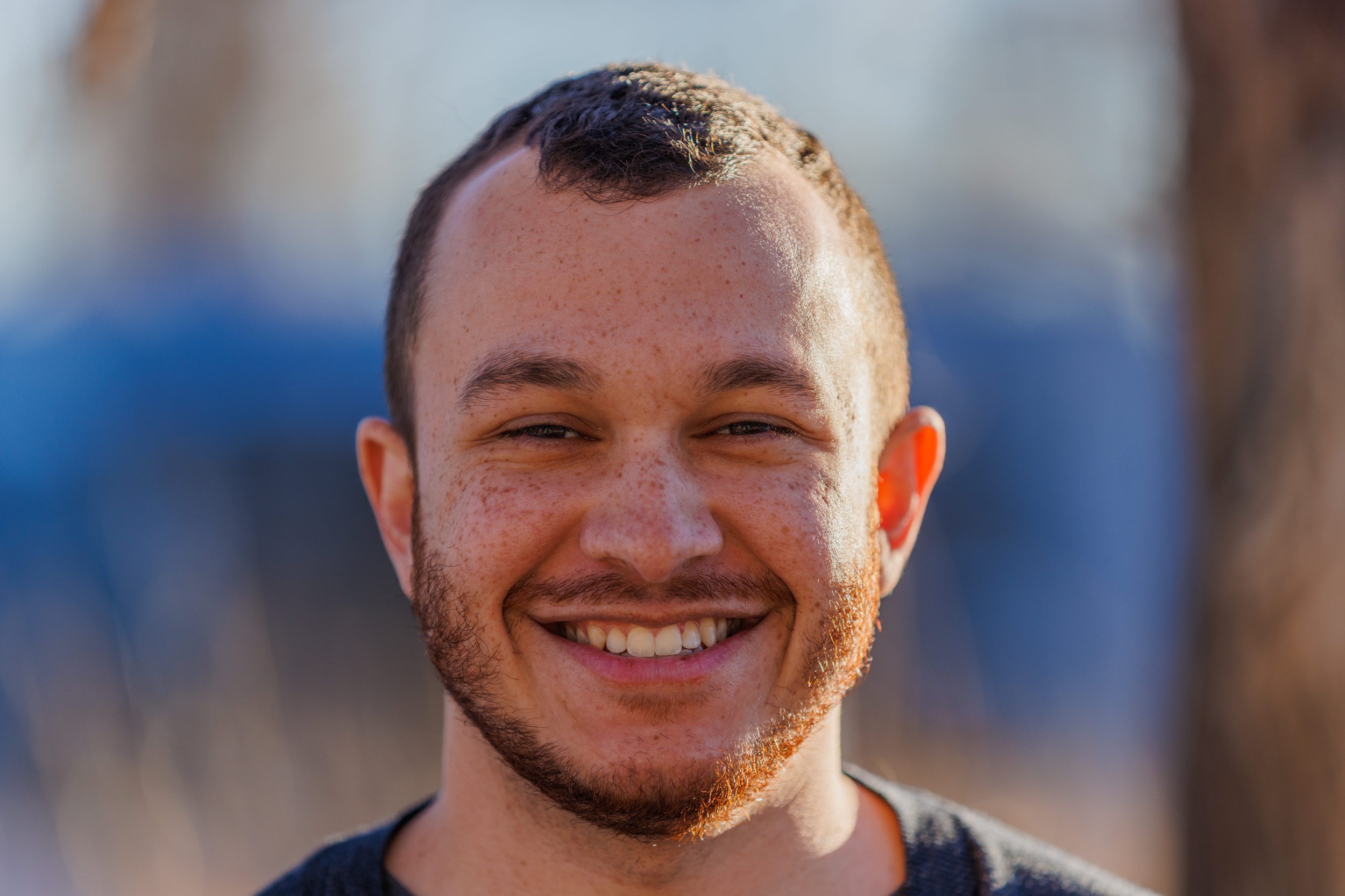 Close-up of a young man's face smiling outdoors, with short brown hair, facial hair, and freckles, in natural sunlight.