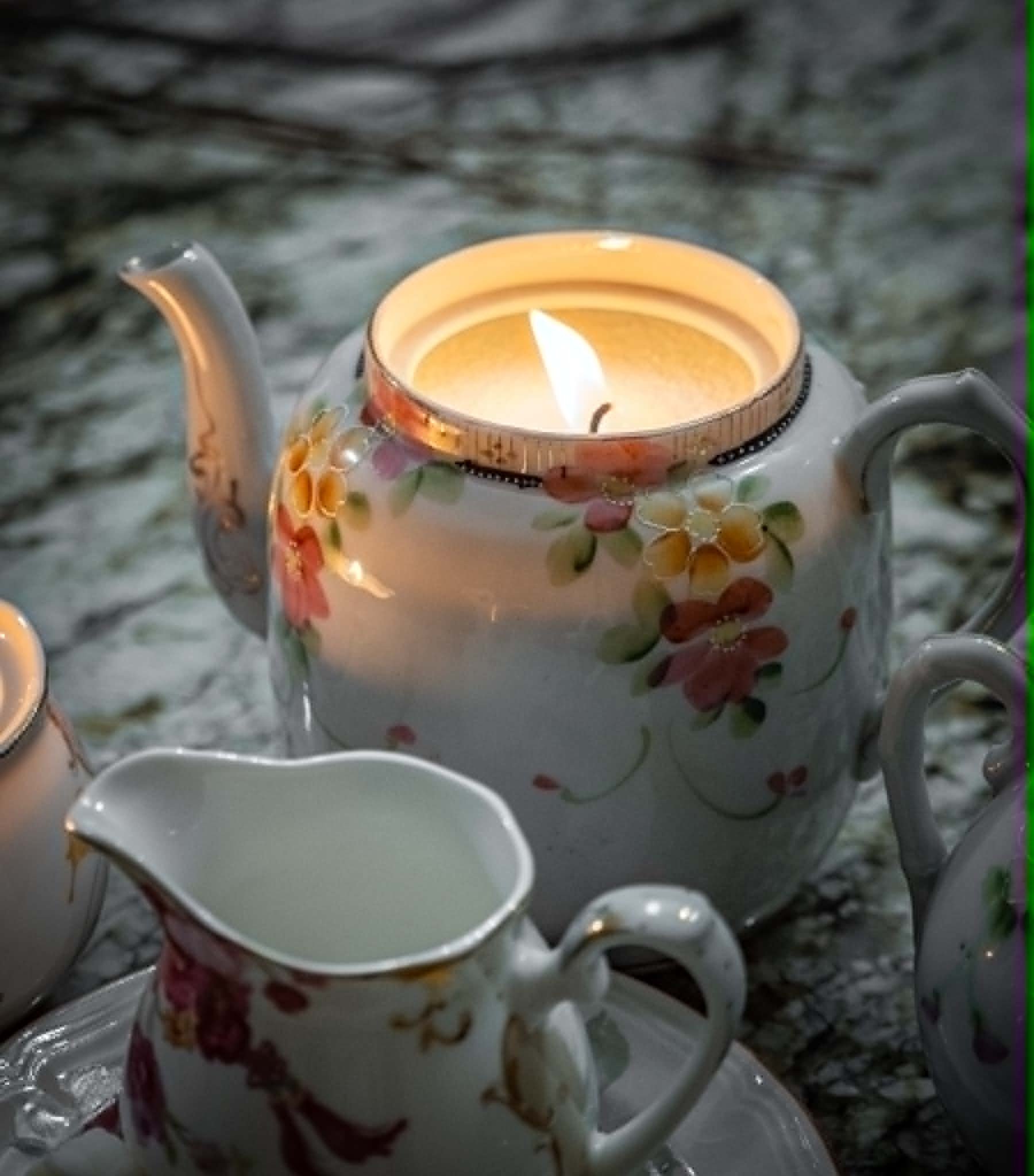 A floral-patterned teapot with a lit candle inside, placed on a reflective surface with small teacups around it.