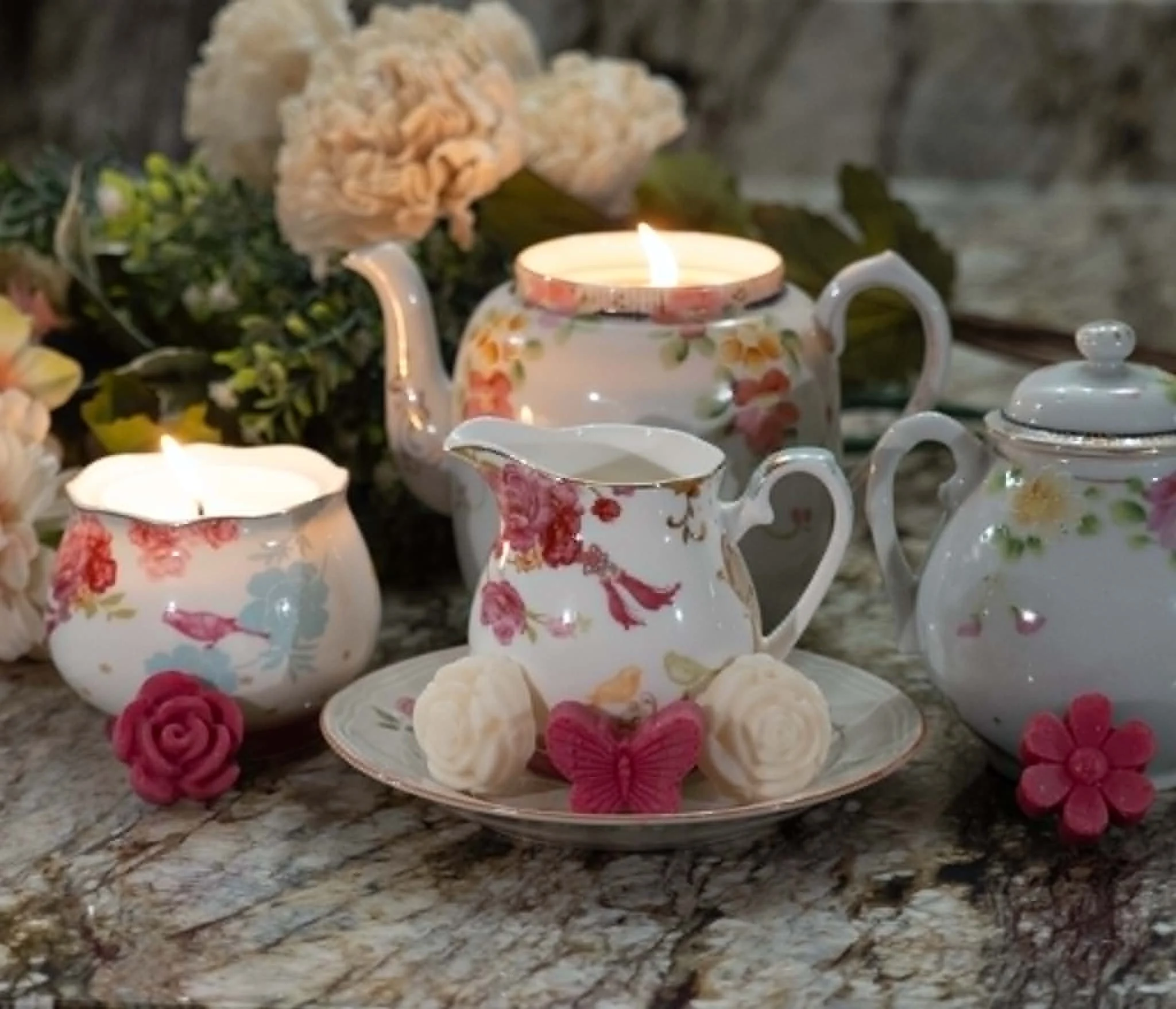 A teapot and cups with a floral design, surrounded by pink roses, set on a table with a flower arrangement and a lit candle in the background.