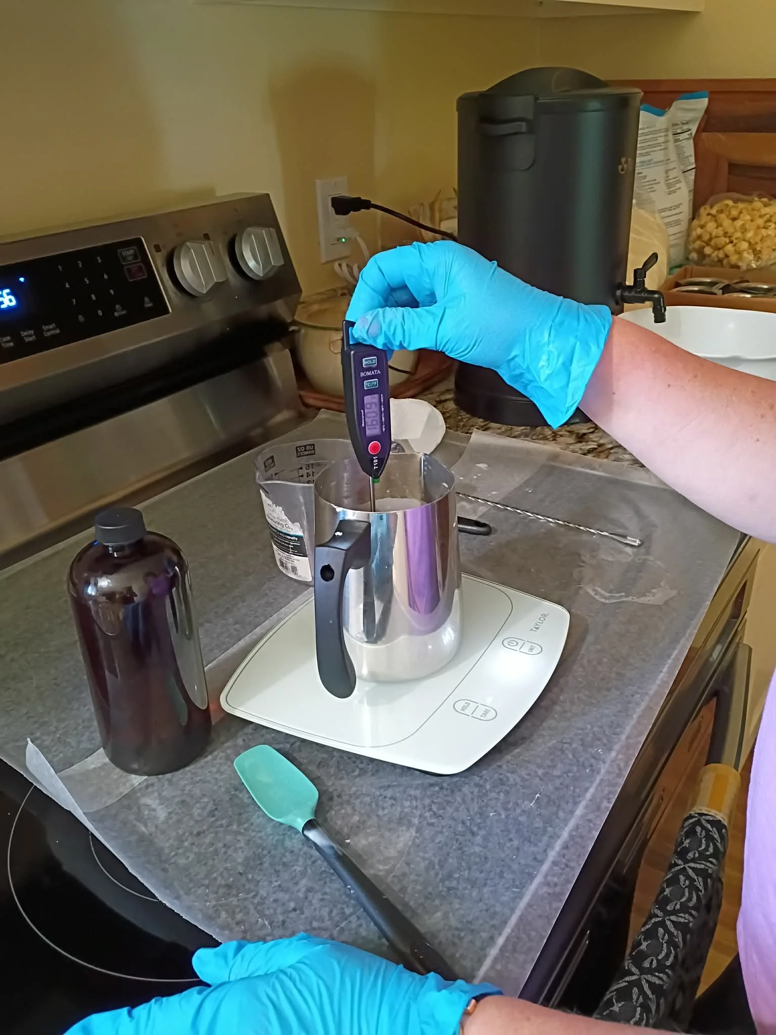 Person in a blue glove using a digital thermometer to check the temperature of a saucepan on a kitchen stovetop.