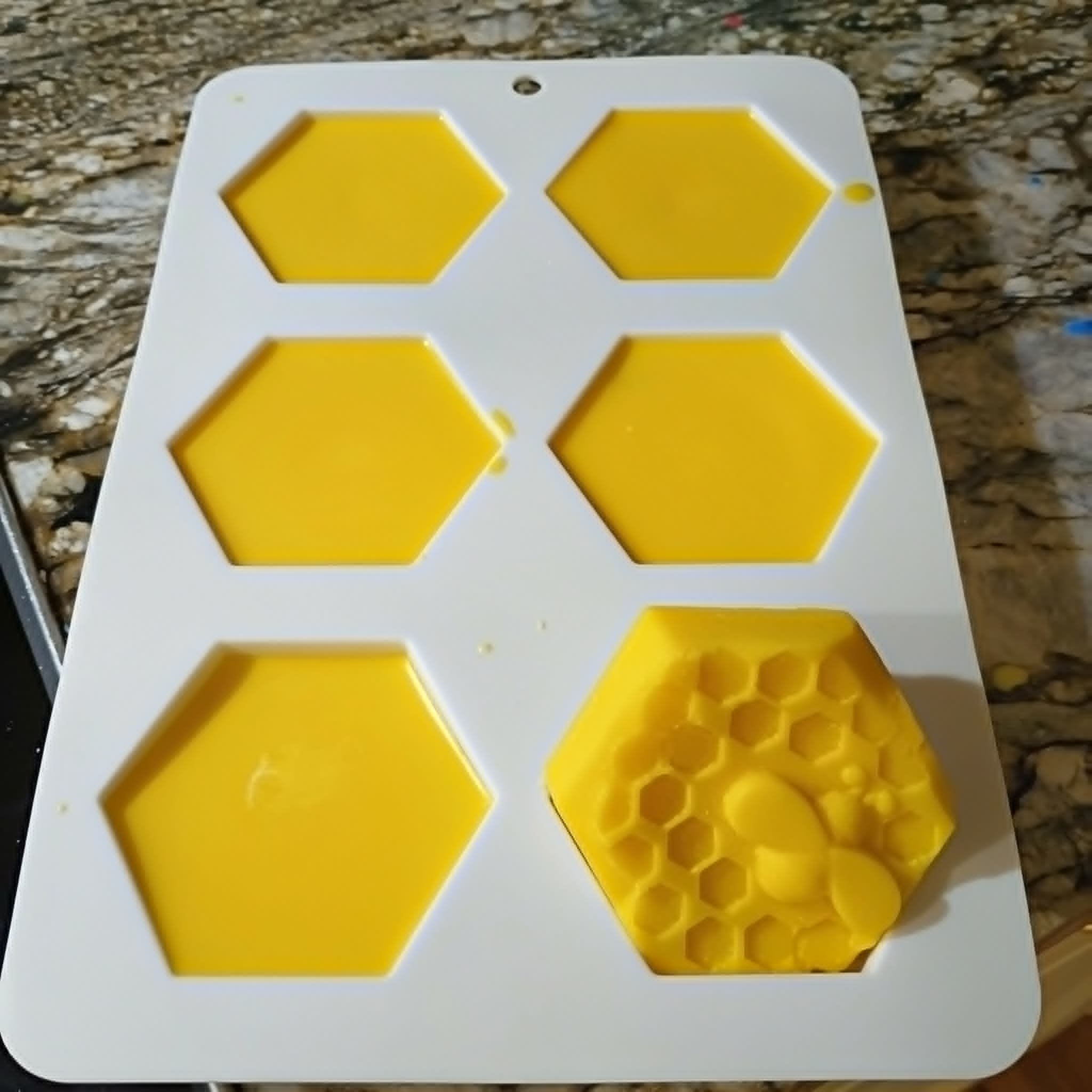 A white plastic device with yellow hexagonal shapes and small yellow compartments, used as a glue mold or casting tray, placed on a wooden surface.