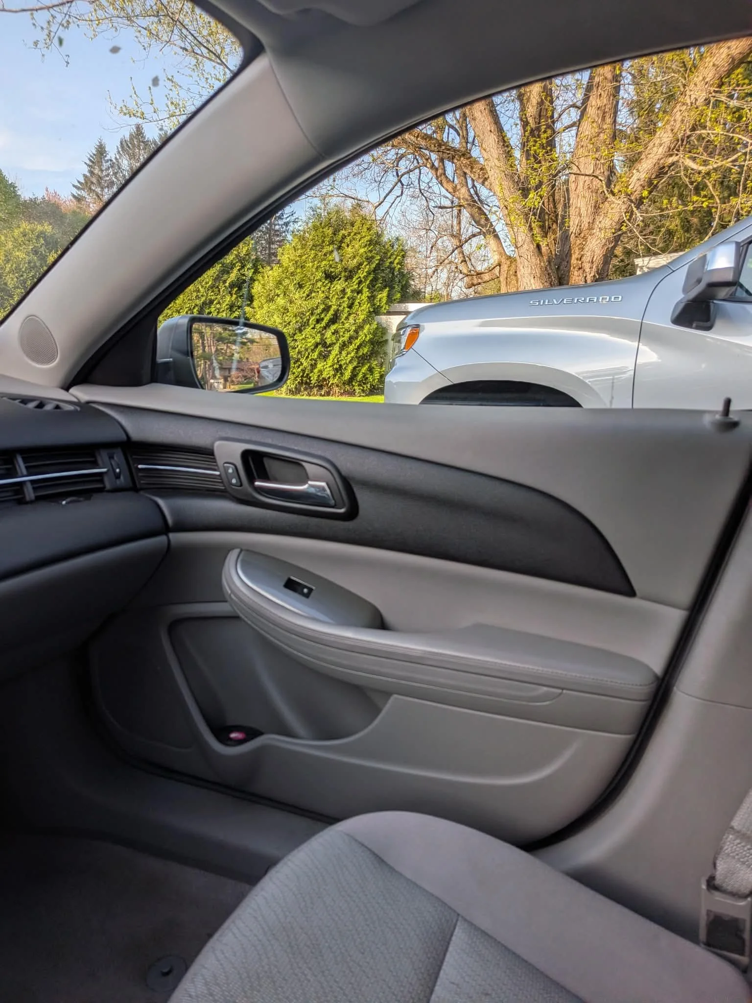 View of the inside of a car from the passenger seat, showing the dashboard, passenger door, and part of the seat. Through the window, a white Chevrolet Silverado truck, trees, and a clear sky are visible.
