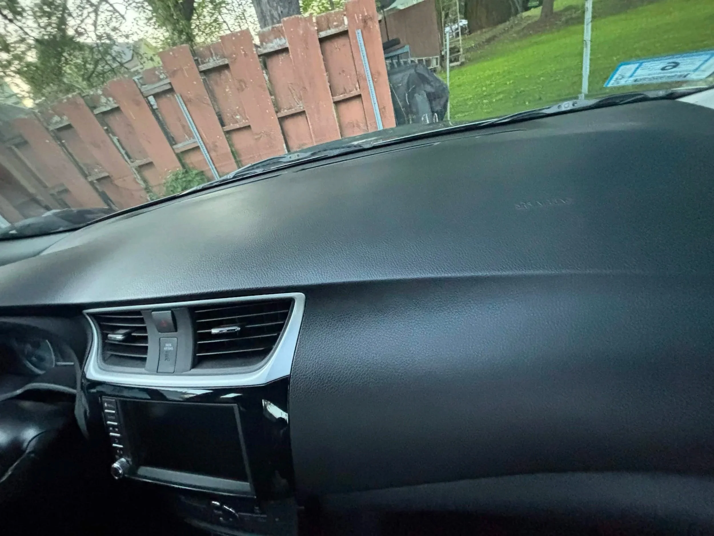 Photo taken from inside a car looking out through the windshield. A wooden fence and a green lawn are visible outside, along with some trees and a cloudy sky.
