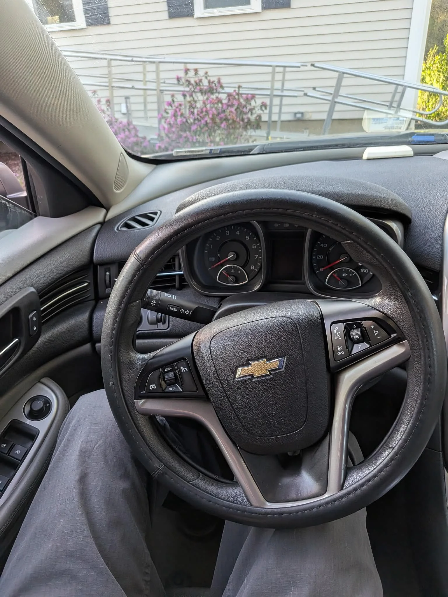 View from inside a car showing the steering wheel with Chevrolet logo, dashboard with gauges, and the front seats. The person is wearing gray pants, and an outside house with siding and pink flowering bushes are visible through the windshield.
