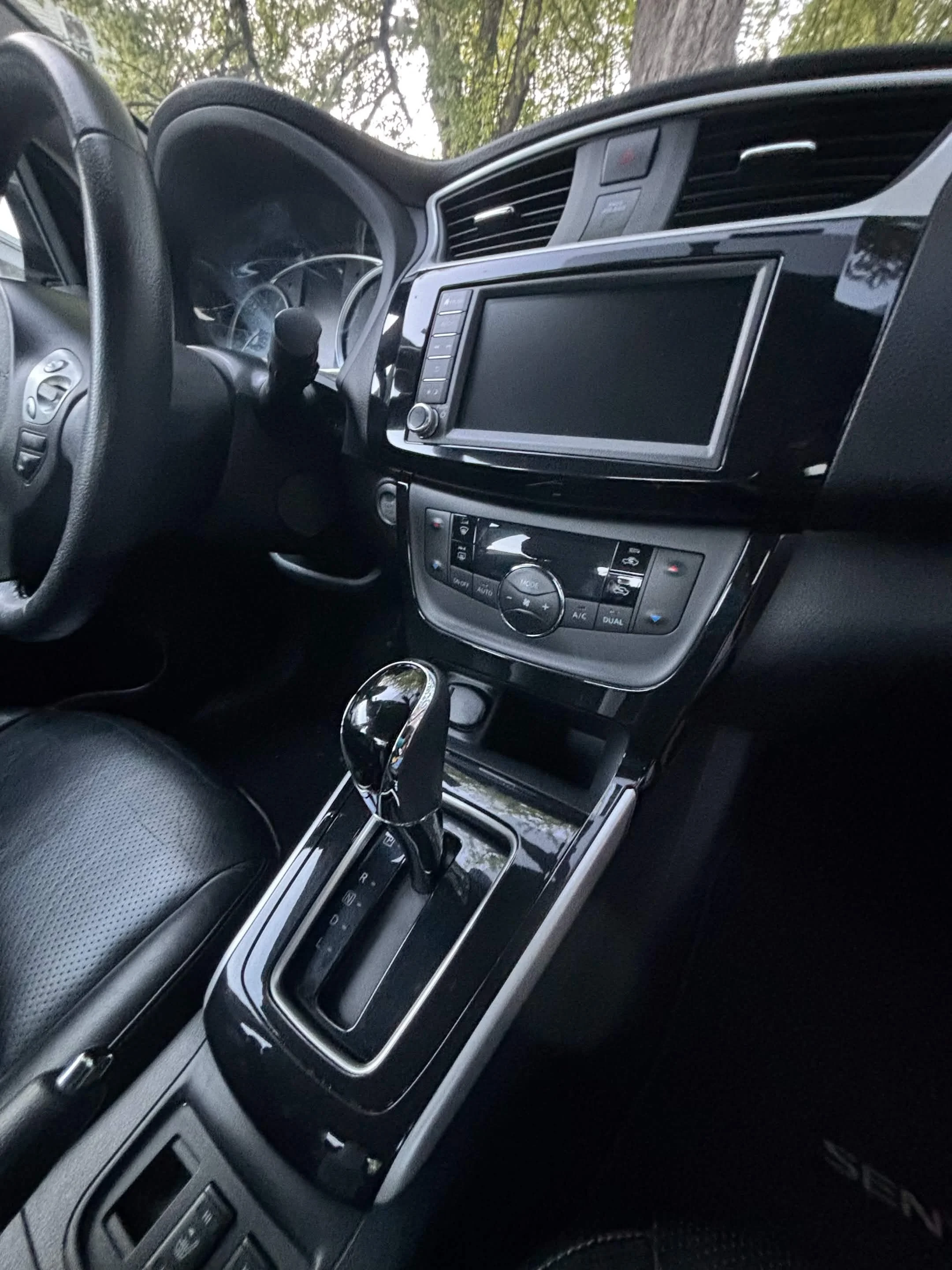 The interior of a car showing the dashboard, steering wheel, center console, gear shift, and part of the front seat with a tree visible outside the window.