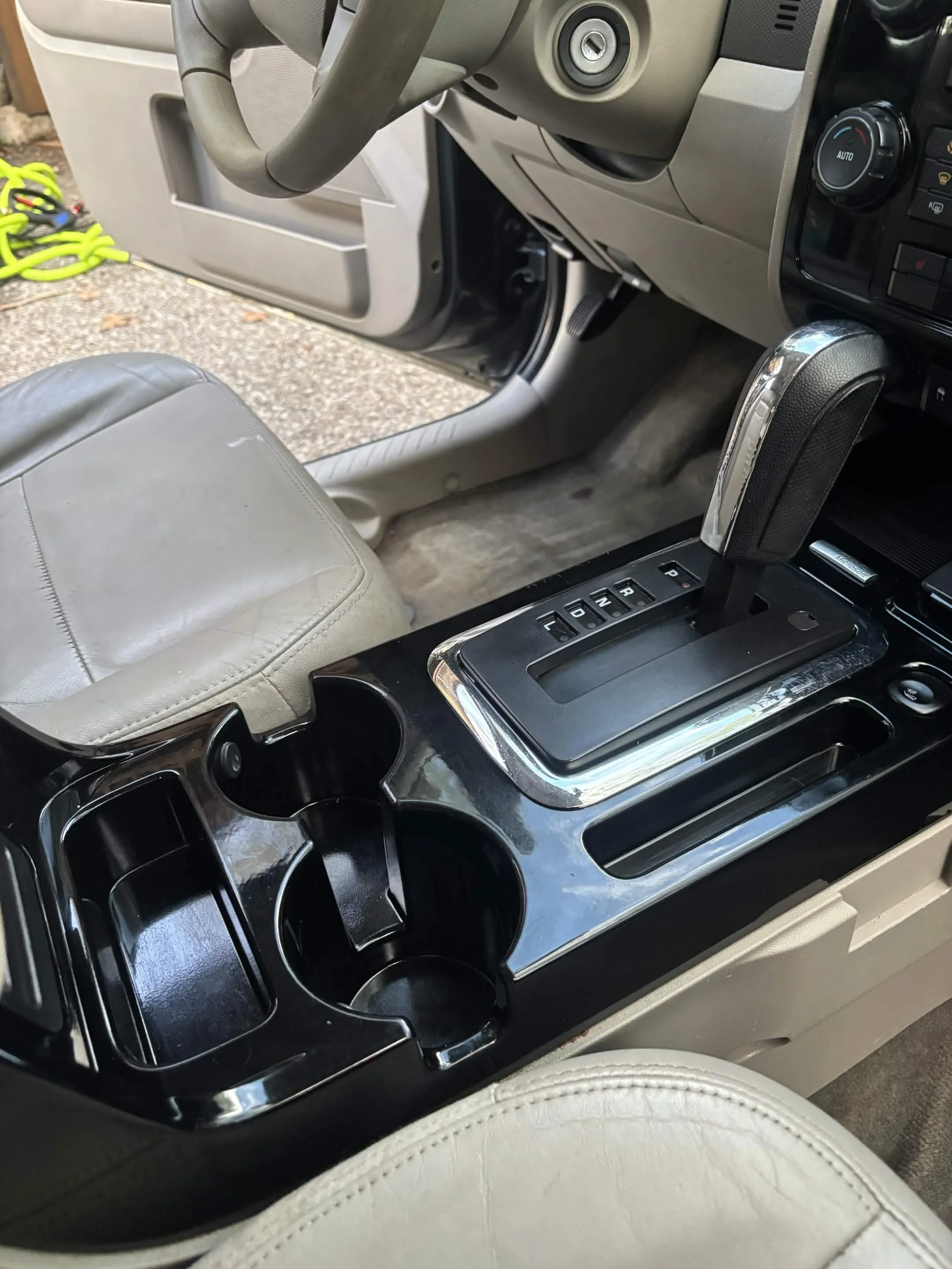 Interior of a vehicle showing the center console with cup holders and gear shift, beige leather seat, and part of the dashboard with climate controls.
