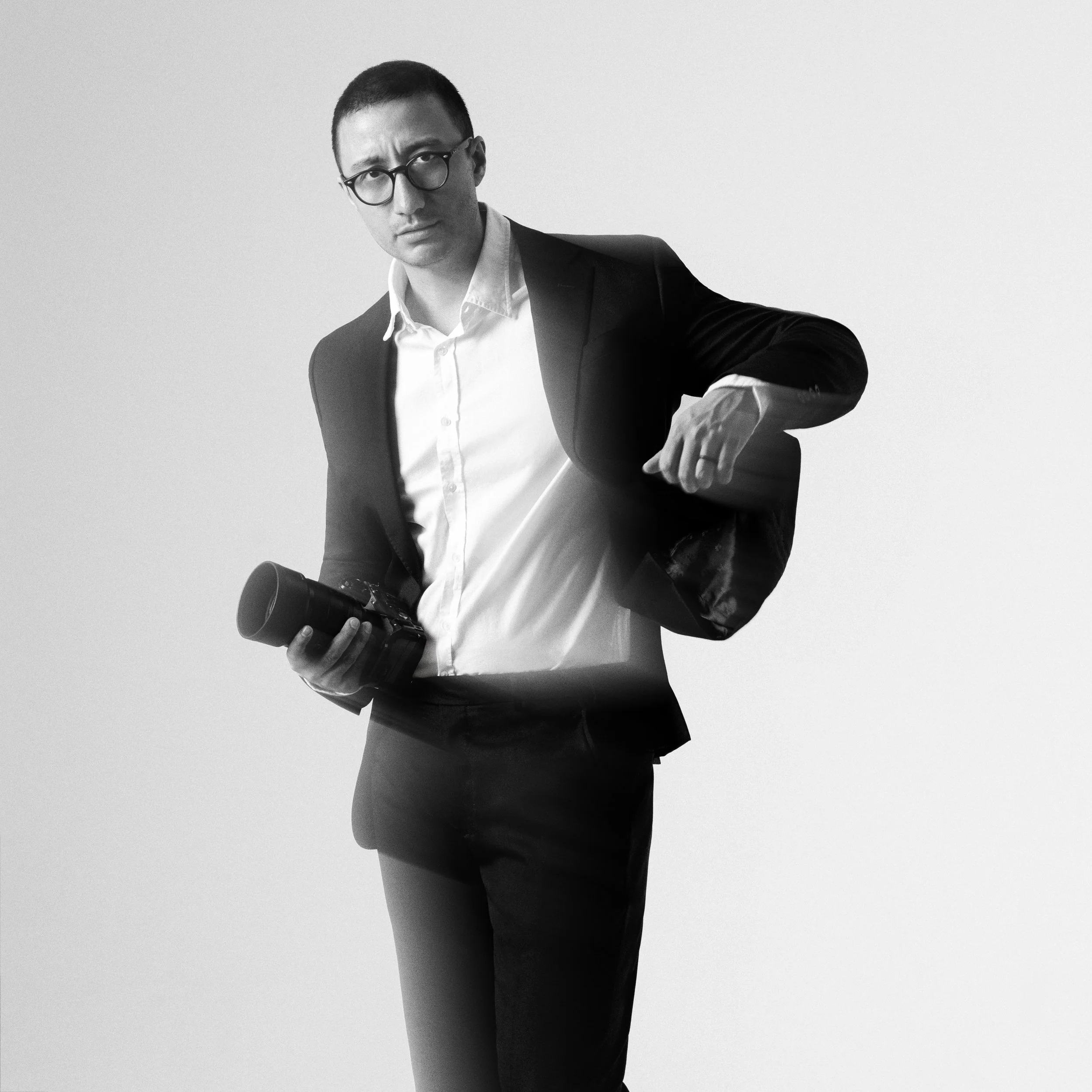 Black and white photo of a man wearing glasses, a white shirt, and a black jacket, holding a camera, with a neutral background.