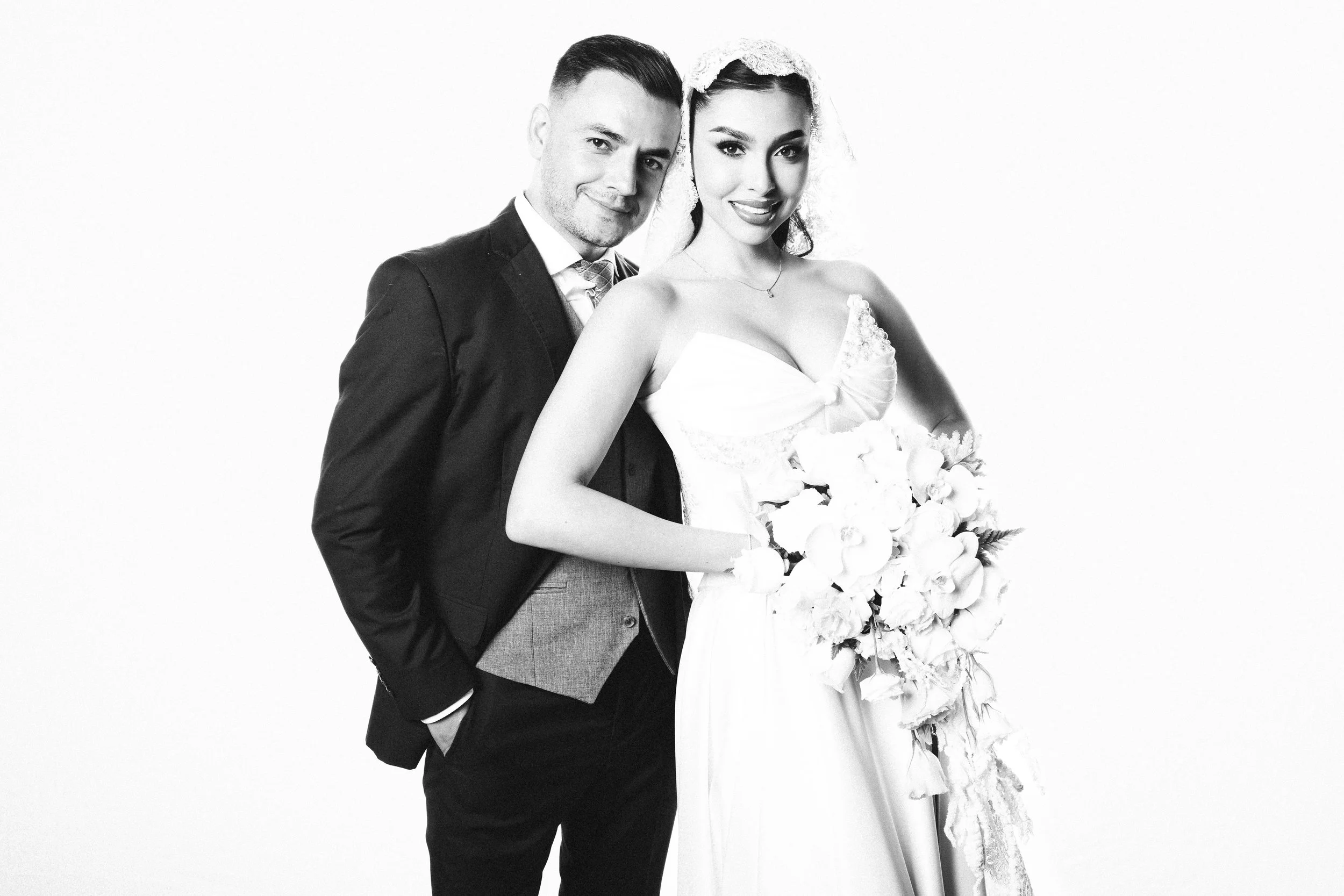 Black and white photo of a newlywed couple, the groom in a suit and tie, the bride in a wedding dress and holding a bouquet of white flowers.
