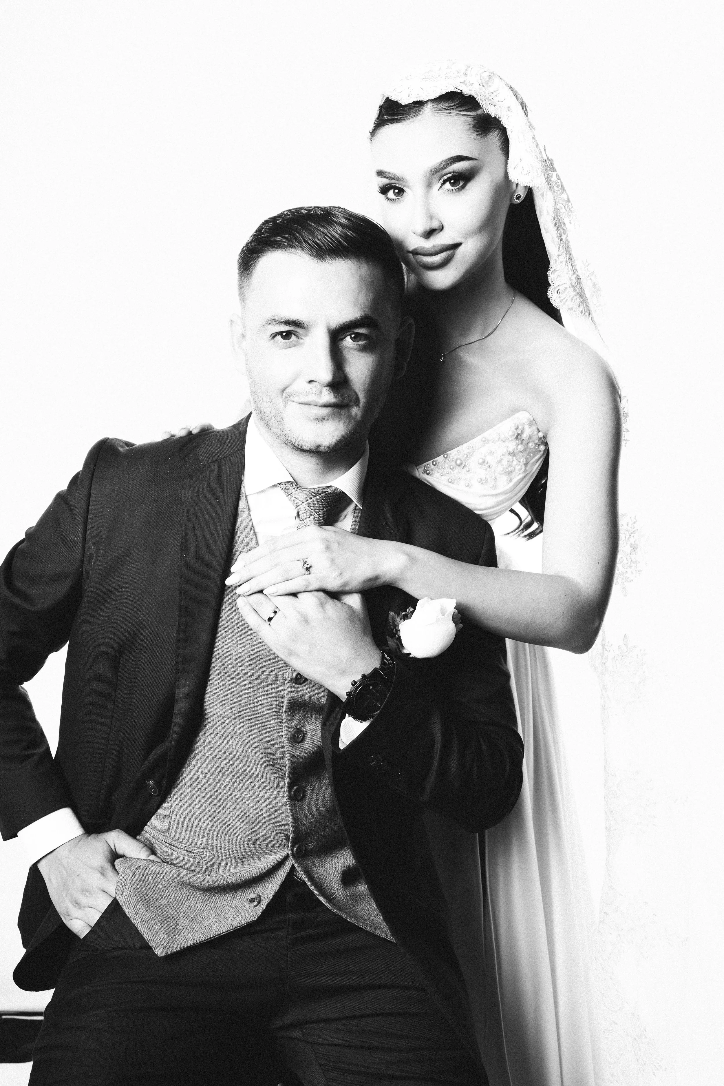 Black and white photo of a man in a suit and a woman in a wedding dress, with the woman standing behind and leaning on the man's shoulder, both smiling, against a plain background.