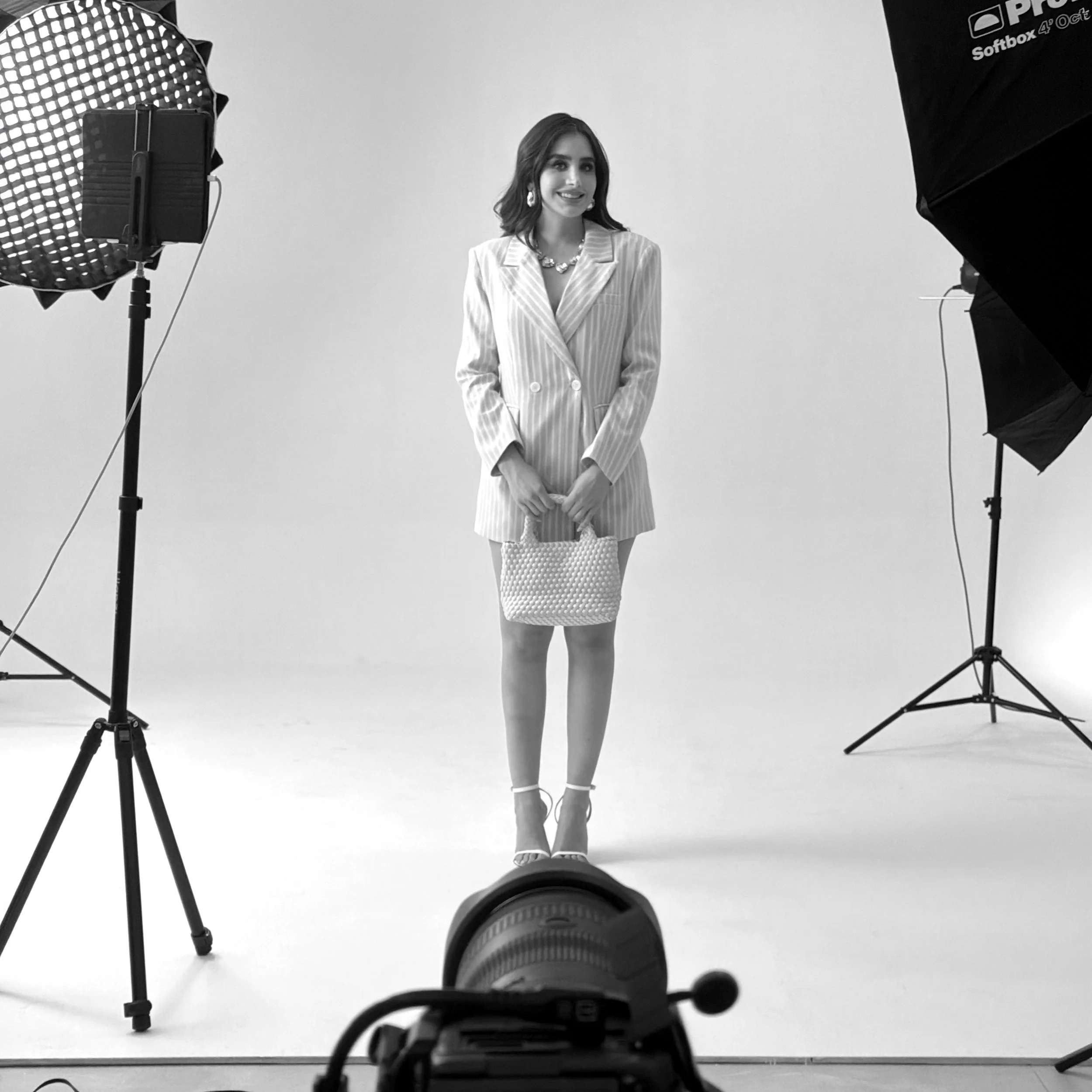 Black and white photo of a woman with shoulder-length hair standing against a plain white background. She is wearing a light-colored, striped blazer, a skirt or dress with a dotted pattern, and high-heeled shoes. Professional studio equipment, including softbox lights and a camera on a tripod, surrounds her, indicating a photoshoot setup.