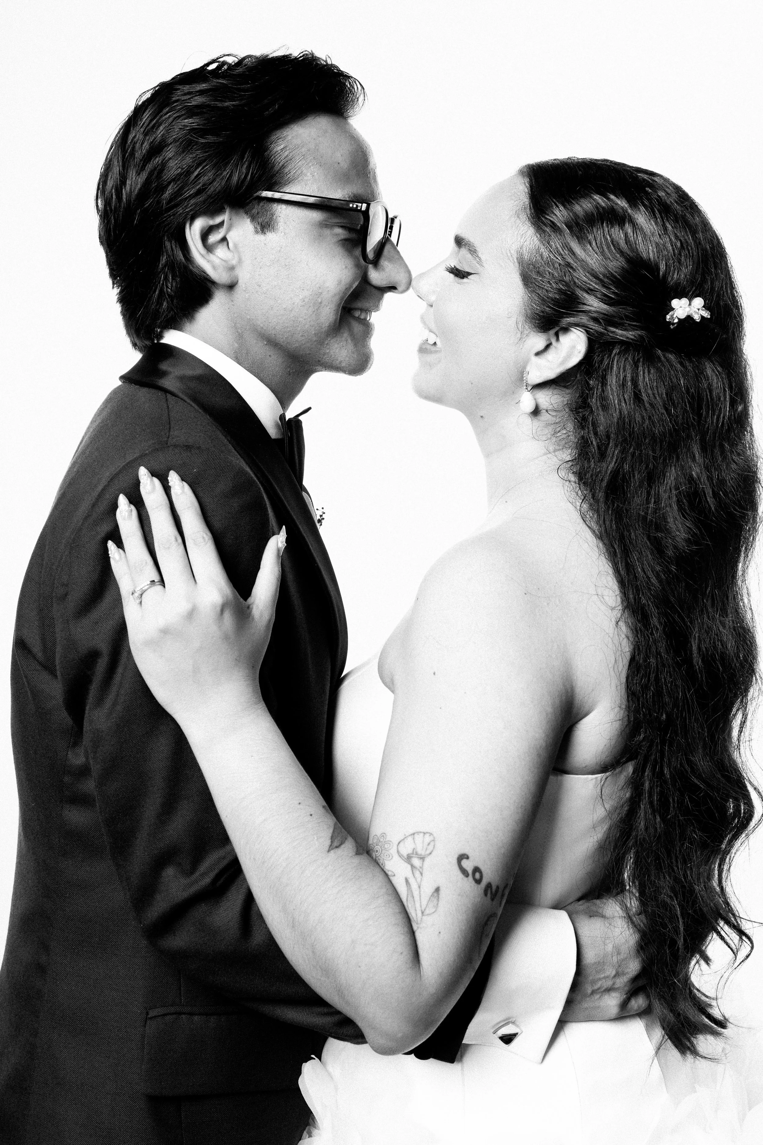 A black and white photo of a couple smiling and about to kiss, with the groom in a tuxedo and the bride in a strapless dress, showing tattoos on her arm.
