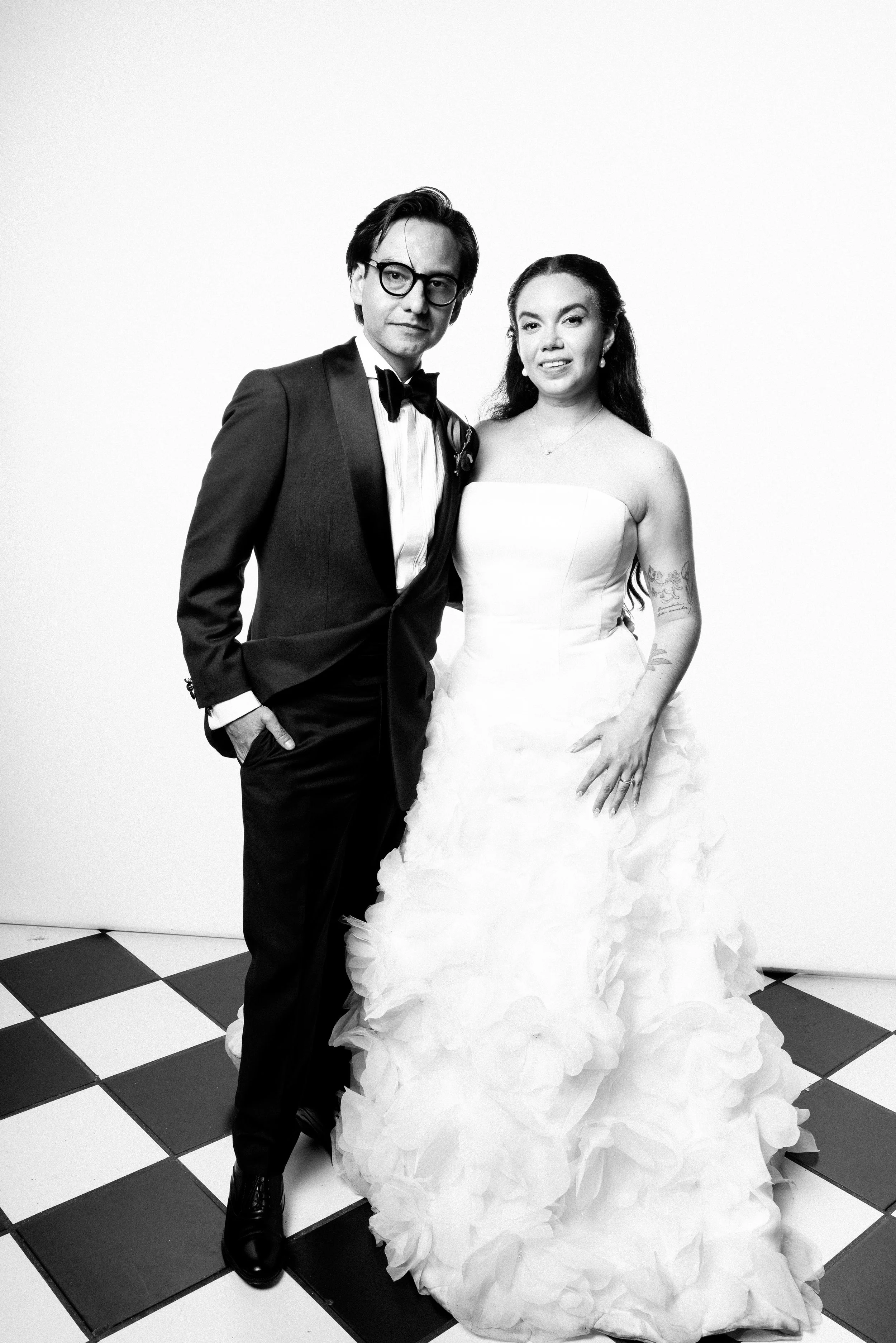 Black and white photo of a man in a tuxedo and a woman in a wedding dress, standing together on a checkered floor. The man has glasses and messy hair, the woman has long wavy hair and tattoos on her arms.