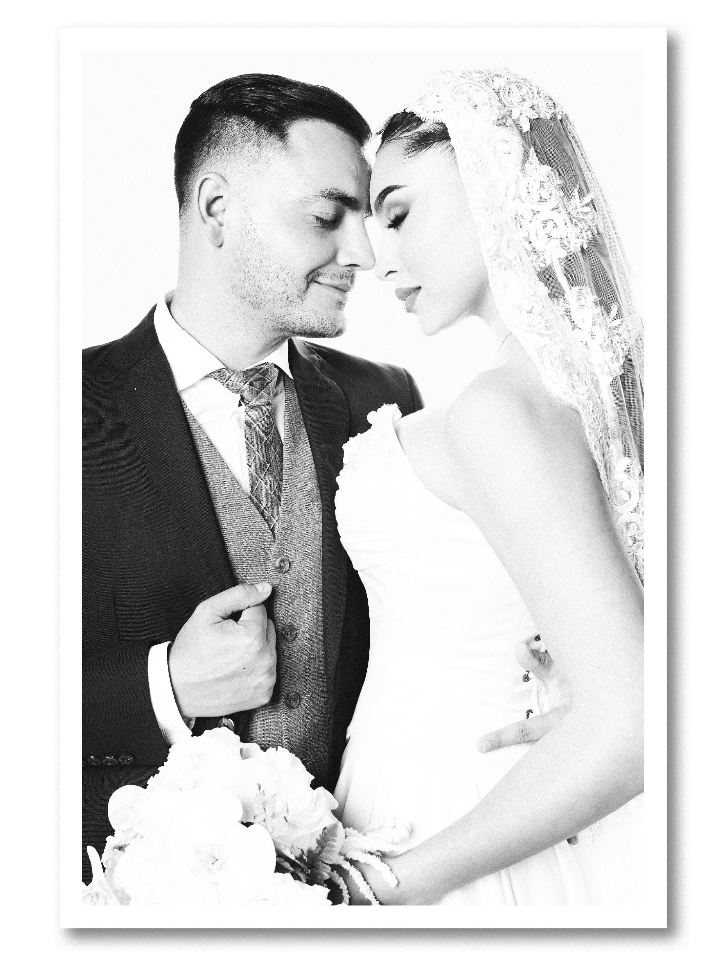 Black and white photo of a wedding couple with their foreheads touching, the groom in a suit and vest and the bride in a lace wedding dress and veil, holding a bouquet of flowers.